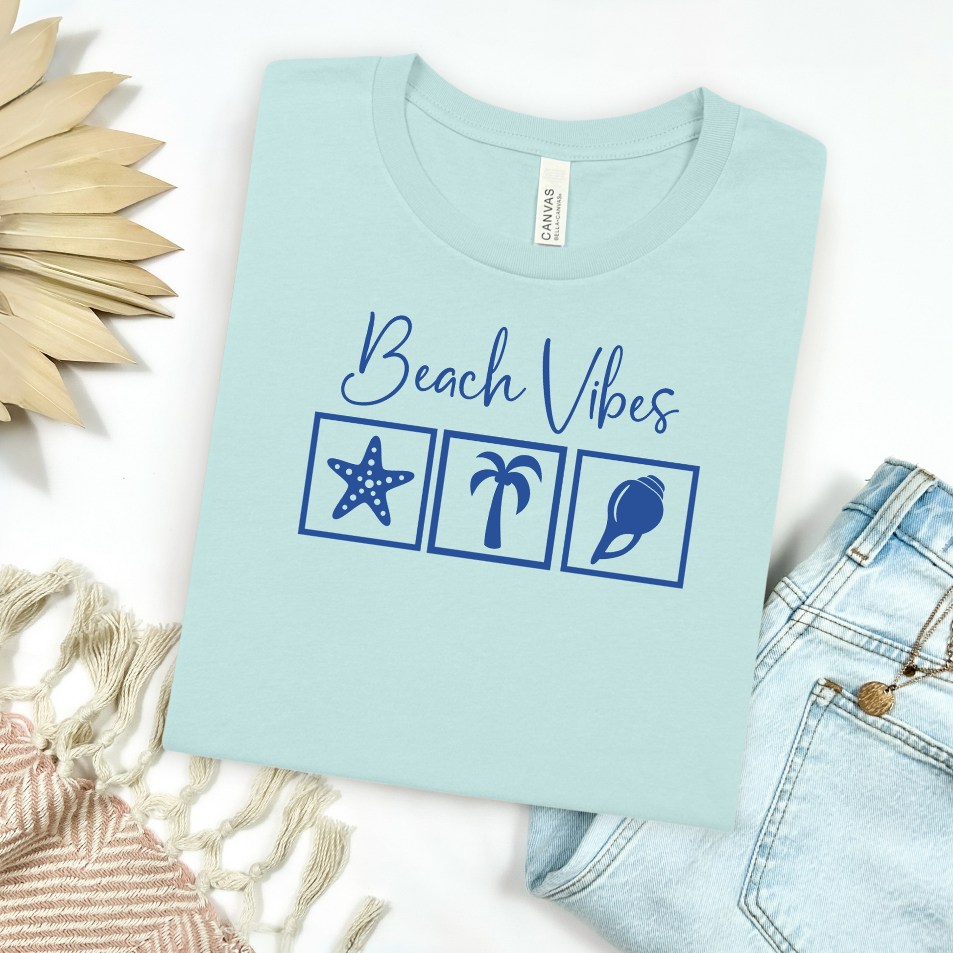 Bella and Canvas 3001 "Beach Vibes" t shirt with a starfish, palm tree, and a seashell graphics in monochrome color. Picture on a flay lay mockup with pair of blue jeans and blanket and a wicker sun shaped accessory.