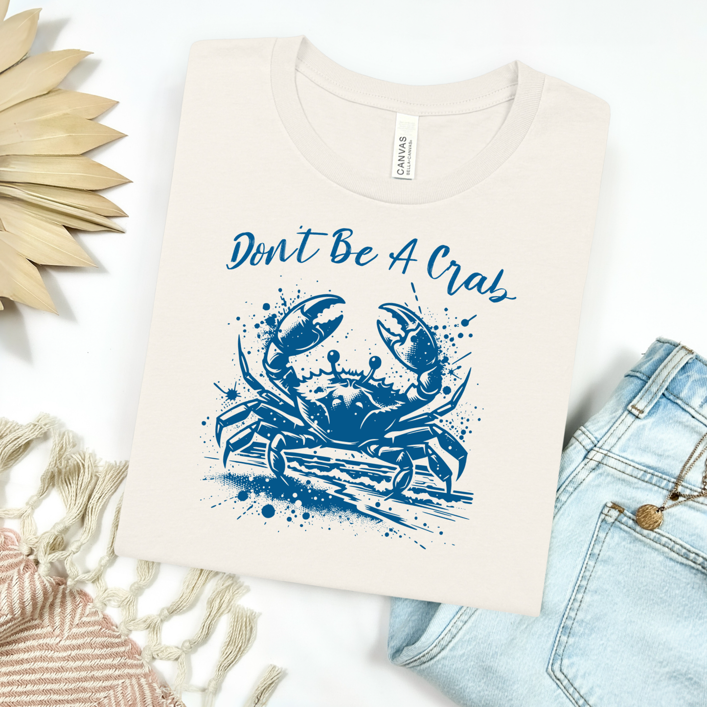 Bella and Canvas 3001 "Don't Be A Crab" t shirt with crab splashing in water one color graphic. Picture on a flay lay mockup with pair of blue jeans and blanket and a wicker sun shaped accessory.