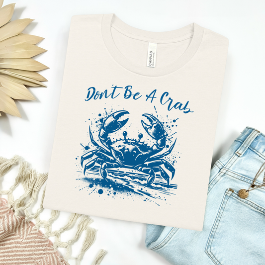 Bella and Canvas 3001 "Don't Be A Crab" t shirt with crab splashing in water one color graphic. Picture on a flay lay mockup with pair of blue jeans and blanket and a wicker sun shaped accessory.