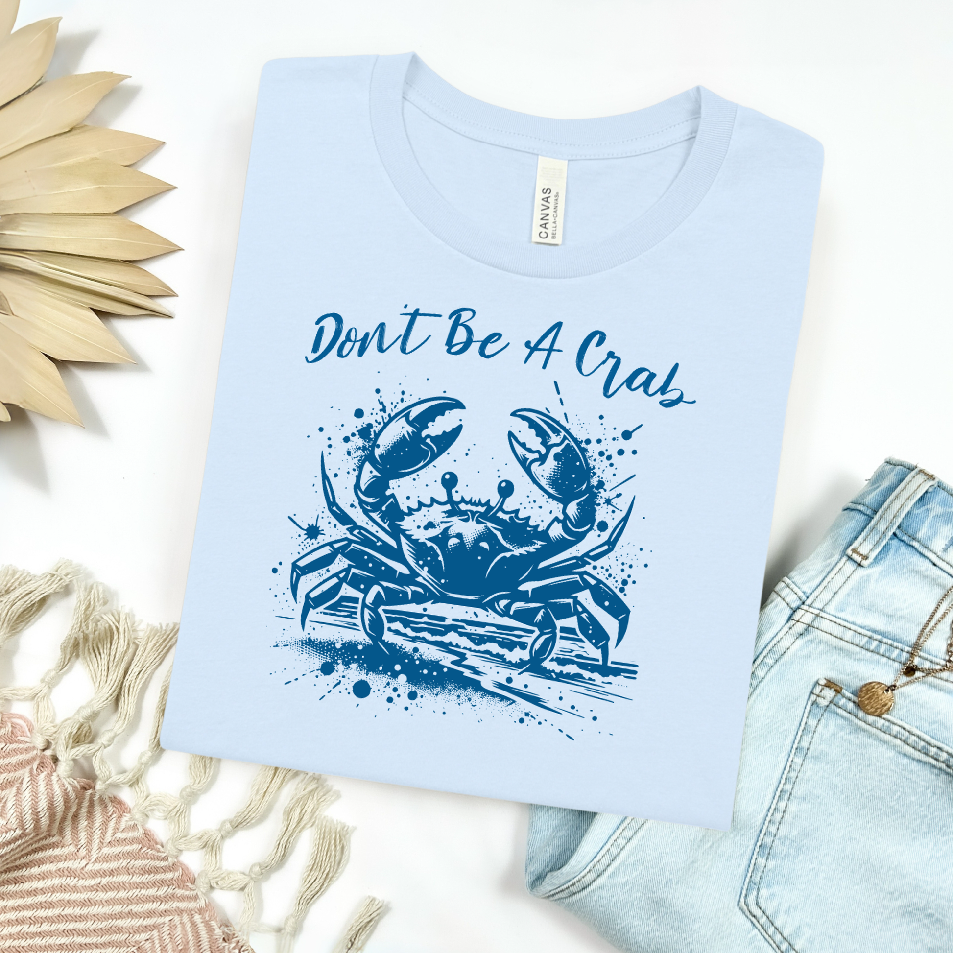 Bella and Canvas 3001 "Don't Be A Crab" t shirt with crab splashing in water one color graphic. Picture on a flay lay mockup with pair of blue jeans and blanket and a wicker sun shaped accessory.