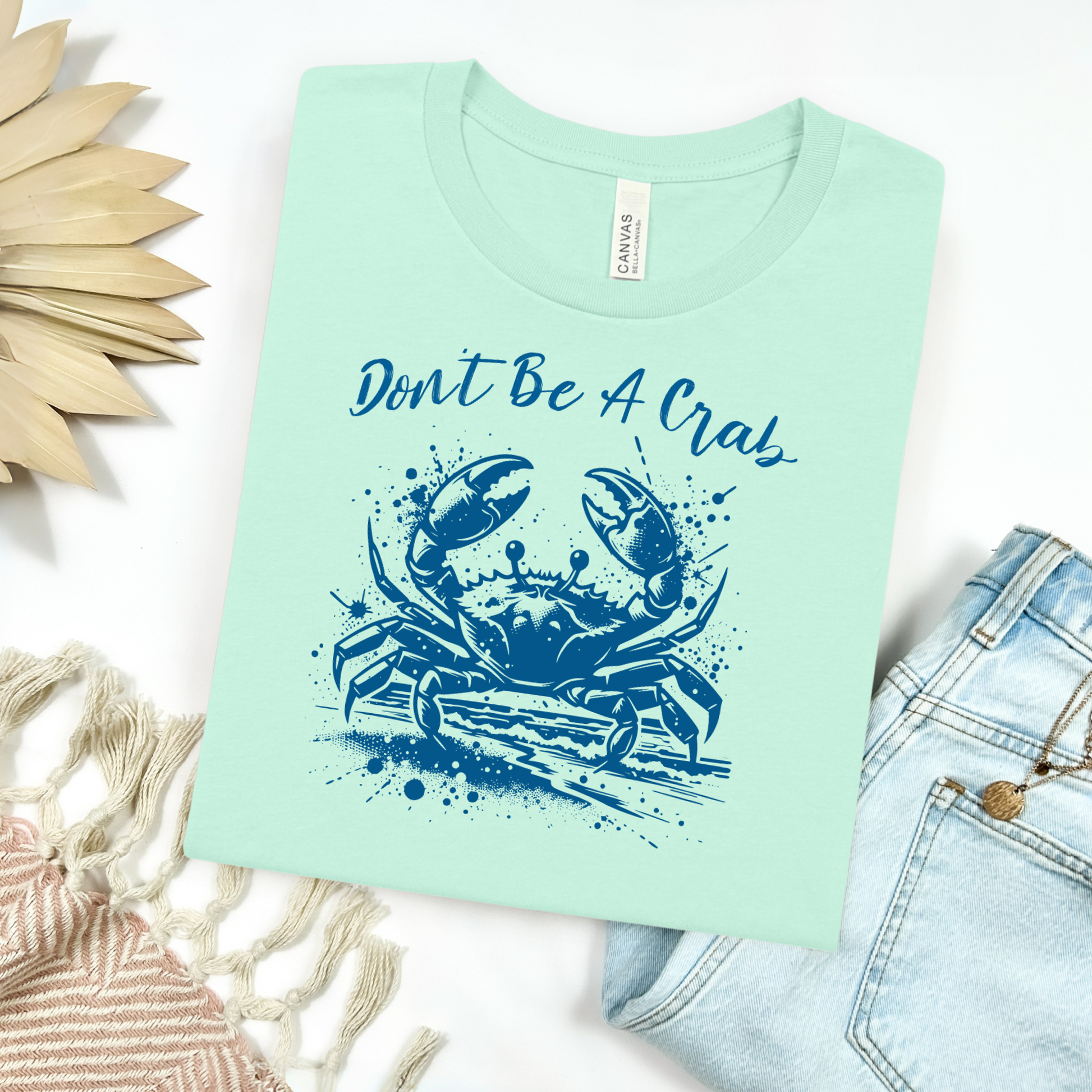 Bella and Canvas 3001 "Don't Be A Crab" t shirt with crab splashing in water one color graphic. Picture on a flay lay mockup with pair of blue jeans and blanket and a wicker sun shaped accessory.