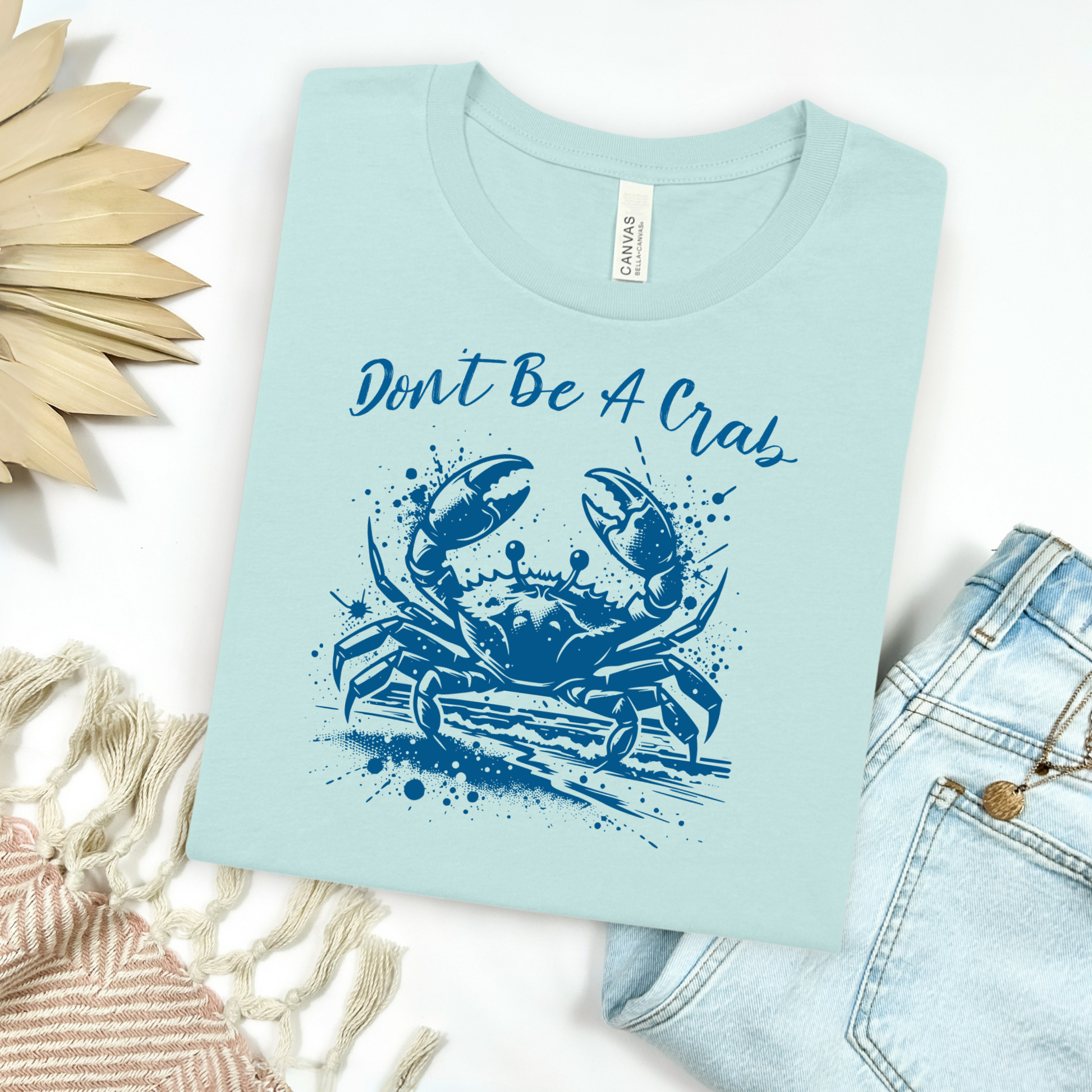 Bella and Canvas 3001 "Don't Be A Crab" t shirt with crab splashing in water one color graphic. Picture on a flay lay mockup with pair of blue jeans and blanket and a wicker sun shaped accessory.