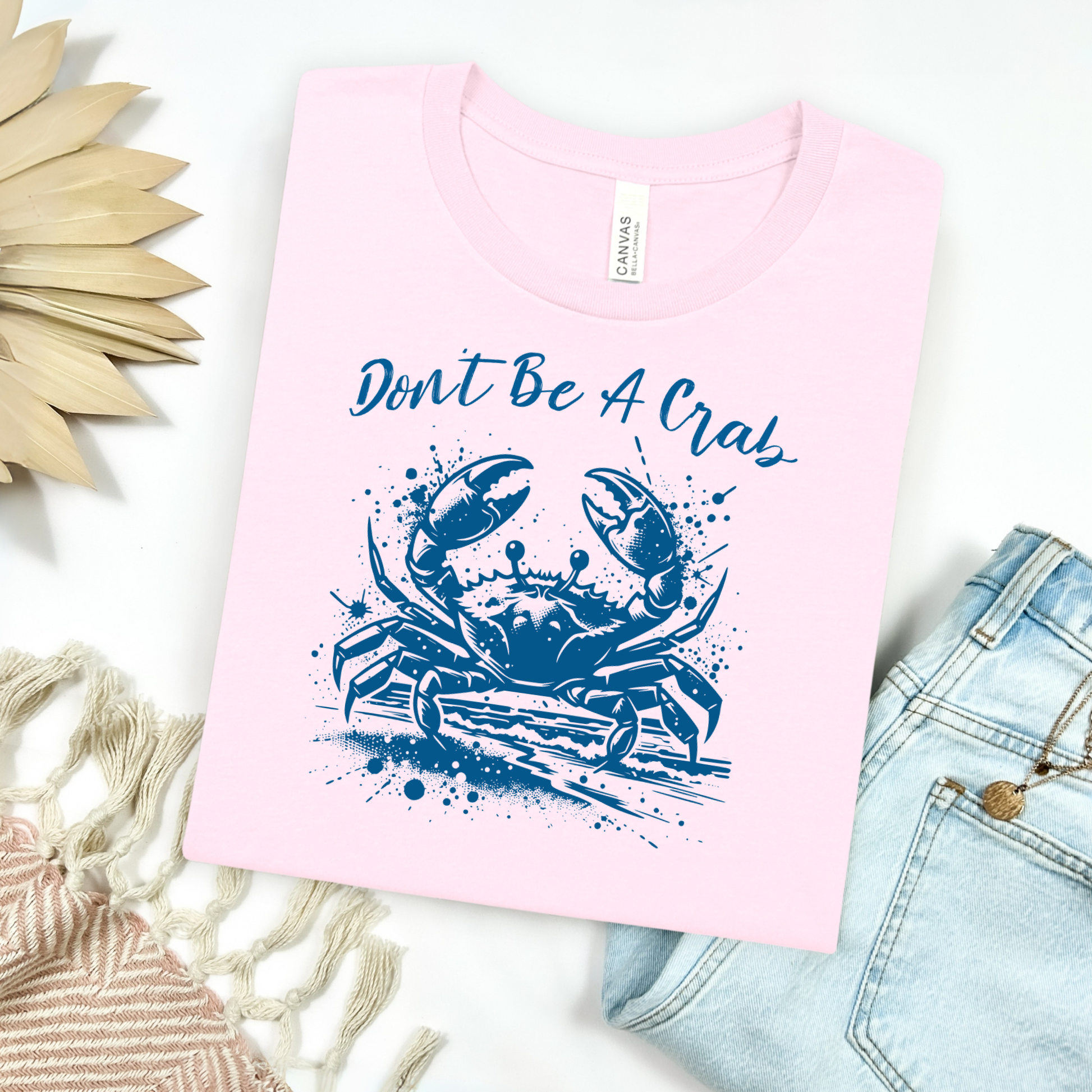 Bella and Canvas 3001 "Don't Be A Crab" t shirt with crab splashing in water one color graphic. Picture on a flay lay mockup with pair of blue jeans and blanket and a wicker sun shaped accessory.