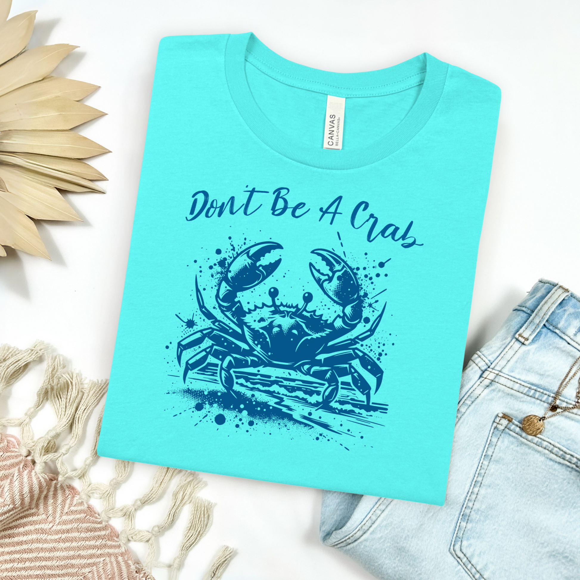 Bella and Canvas 3001 "Don't Be A Crab" t shirt with crab splashing in water one color graphic. Picture on a flay lay mockup with pair of blue jeans and blanket and a wicker sun shaped accessory.