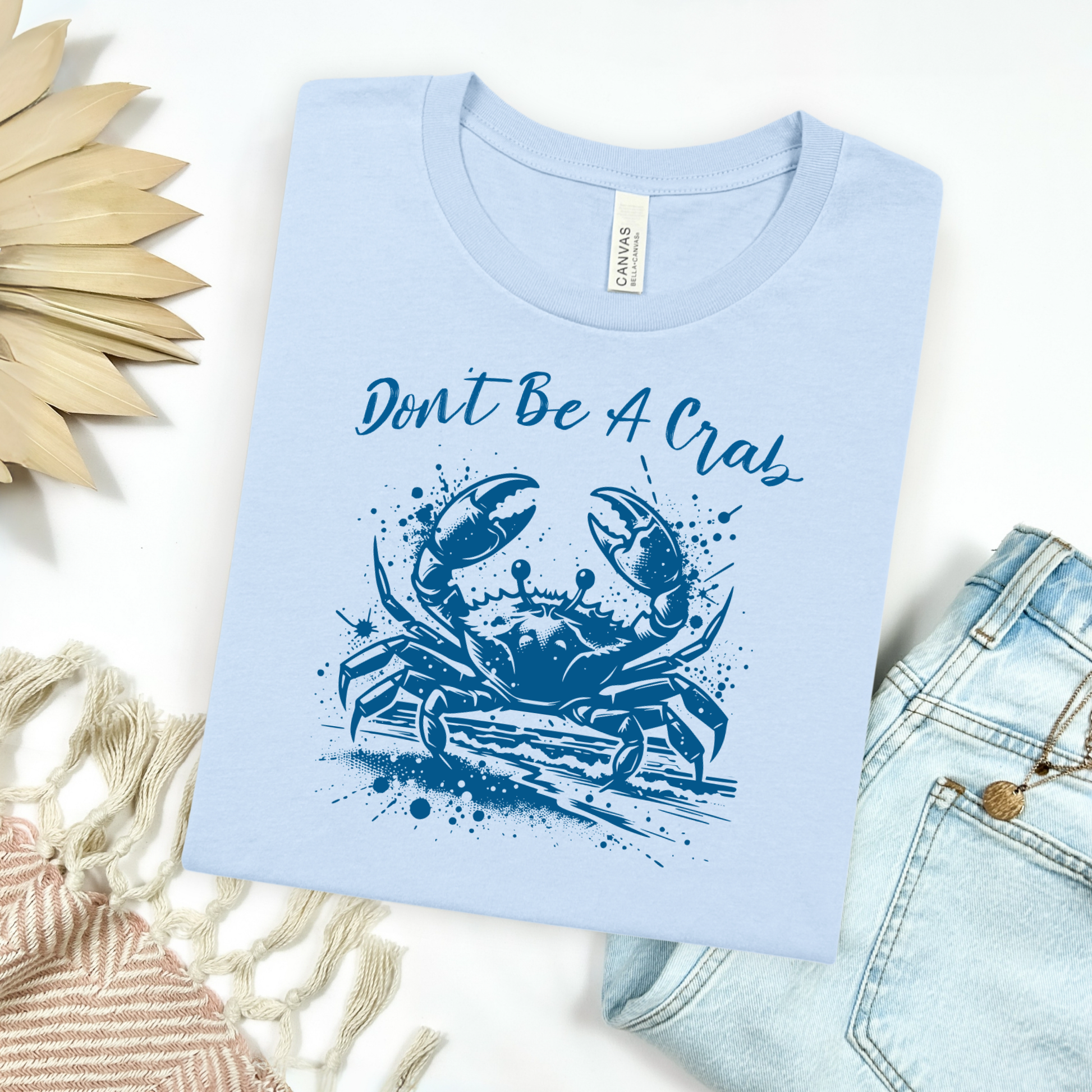 Bella and Canvas 3001 "Don't Be A Crab" t shirt with crab splashing in water one color graphic. Picture on a flay lay mockup with pair of blue jeans and blanket and a wicker sun shaped accessory.