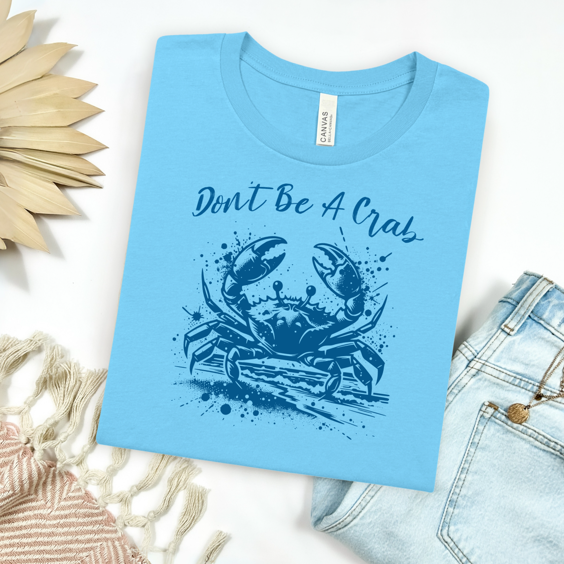 Bella and Canvas 3001 "Don't Be A Crab" t shirt with crab splashing in water one color graphic. Picture on a flay lay mockup with pair of blue jeans and blanket and a wicker sun shaped accessory.
