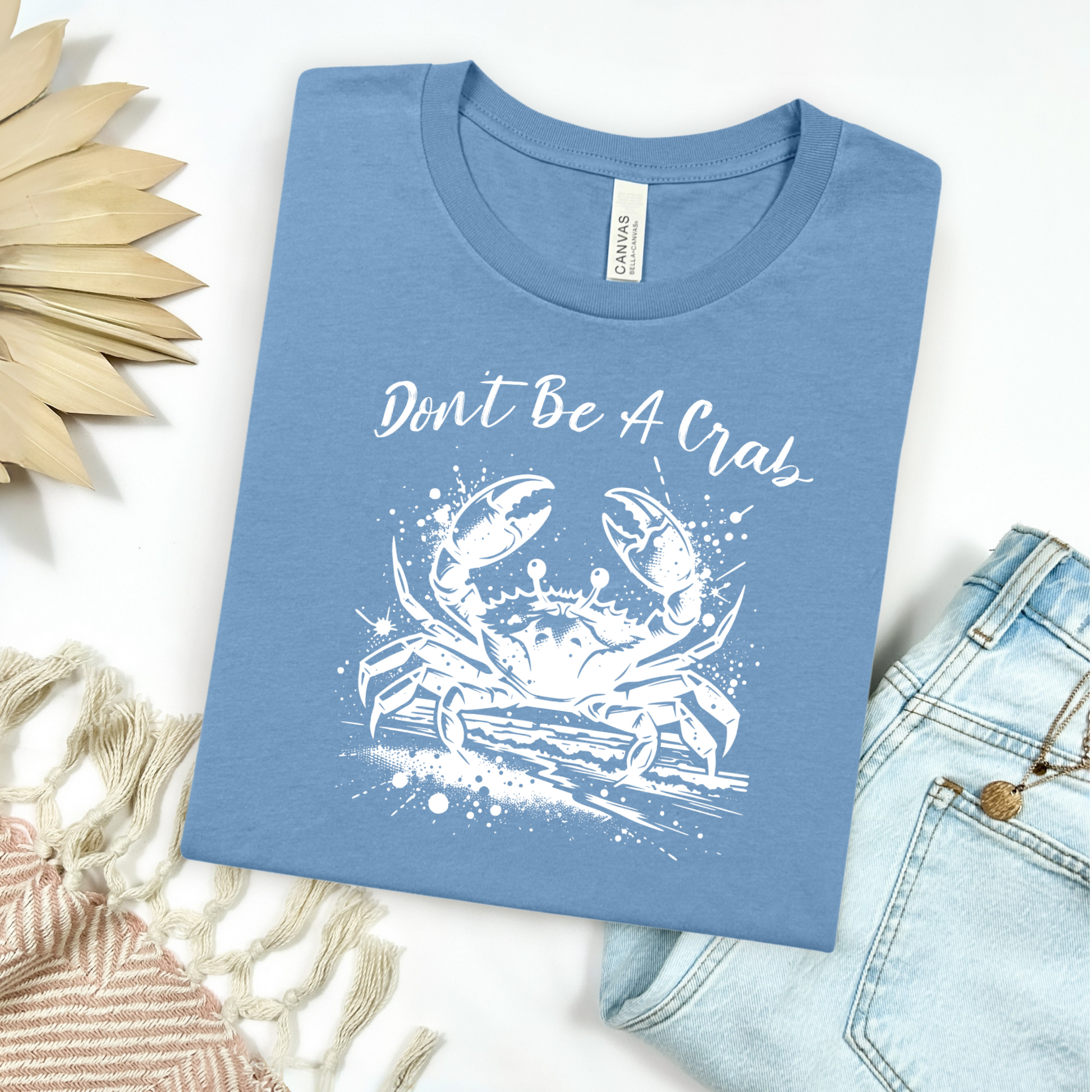 Bella and Canvas 3001 "Don't Be A Crab" t shirt with crab splashing in water one color graphic. Picture on a flay lay mockup with pair of blue jeans and blanket and a wicker sun shaped accessory.