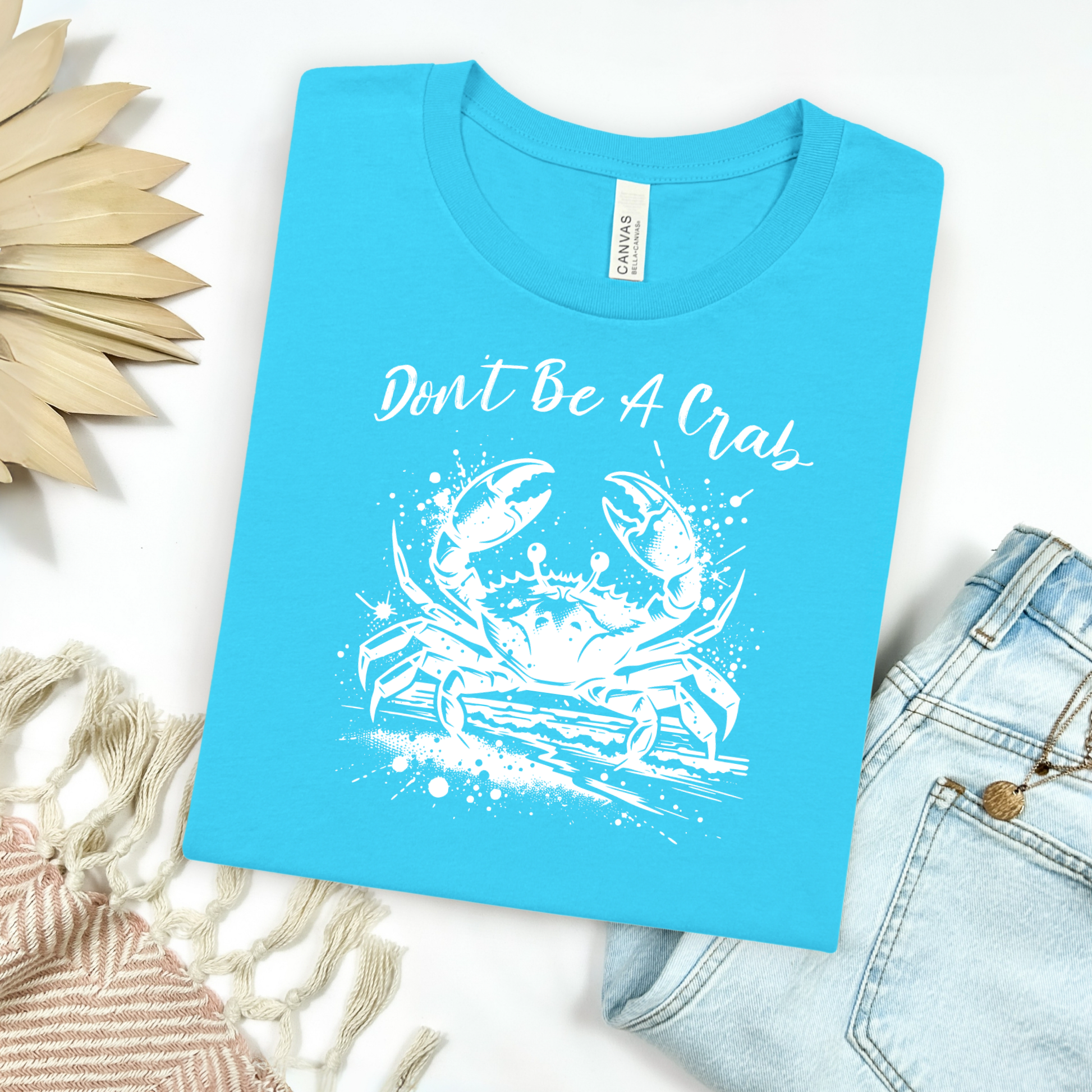 Bella and Canvas 3001 "Don't Be A Crab" t shirt with crab splashing in water one color graphic. Picture on a flay lay mockup with pair of blue jeans and blanket and a wicker sun shaped accessory.