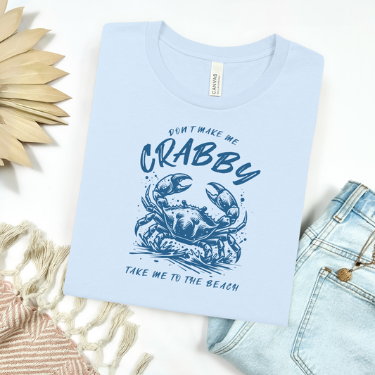 Bella Canvas 3001 "Don't Make Me Crabby Take Me To The Beach" t shirt with a crab jumping in water in a one color graphic. Picture on a flay lay mockup with pair of blue jeans and blanket and a wicker sun shaped accessory.