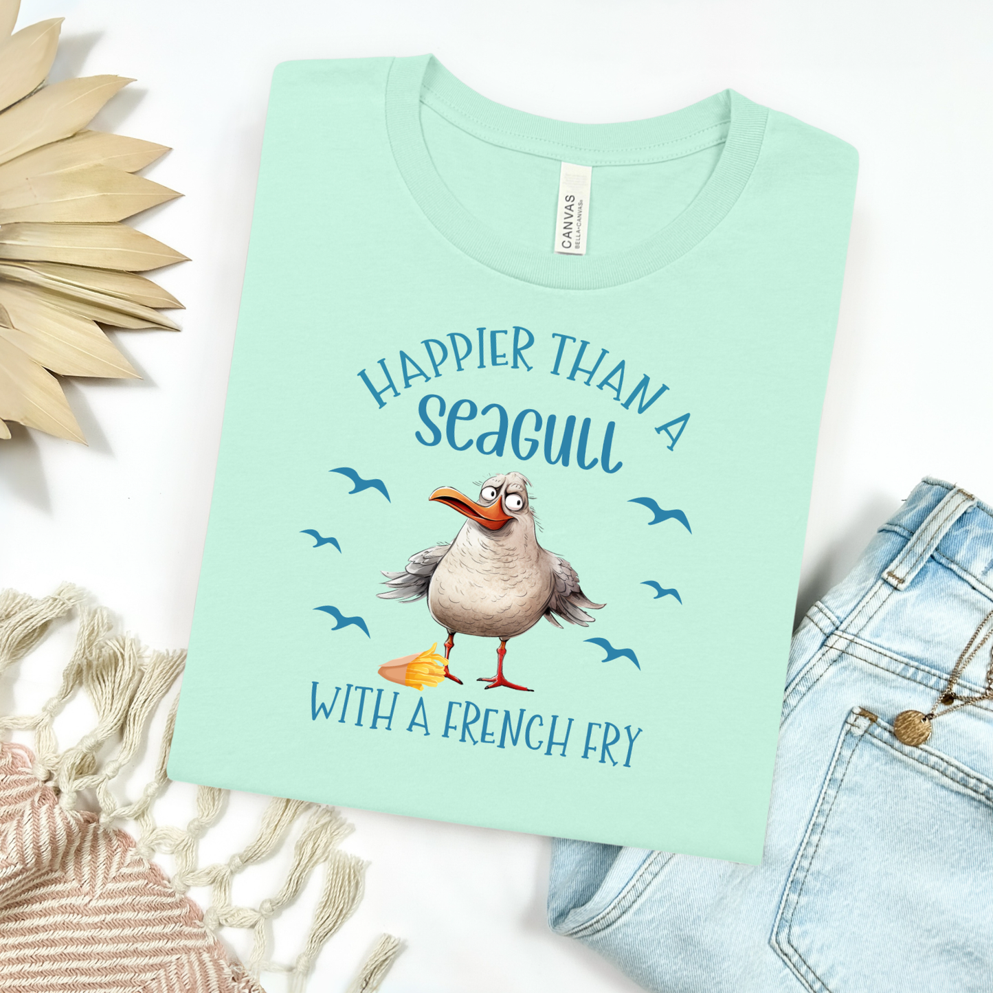 Bella and Canvas 3001 "Happier Than A Seagull With A French Fry" t shirt with an illustration of a seagull with a box of French fries and seagulls flying in the air. Picture on a flay lay mockup with pair of blue jeans and blanket and a wicker sun shaped accessory.