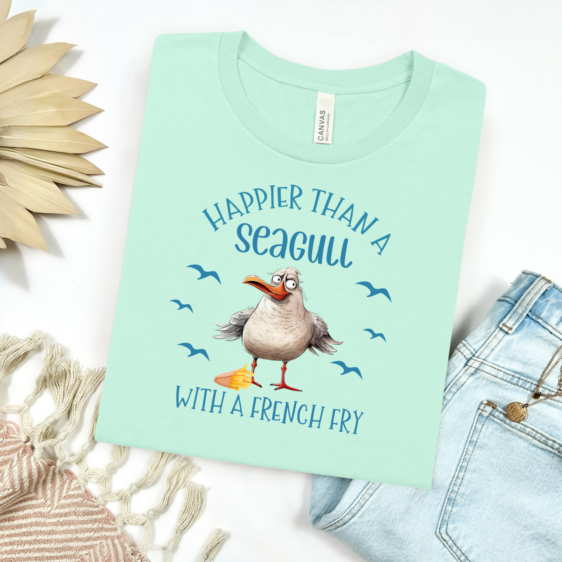 Bella and Canvas 3001 "Happier Than A Seagull With A French Fry" t shirt with an illustration of a seagull with a box of French fries and seagulls flying in the air. Picture on a flay lay mockup with pair of blue jeans and blanket and a wicker sun shaped accessory.