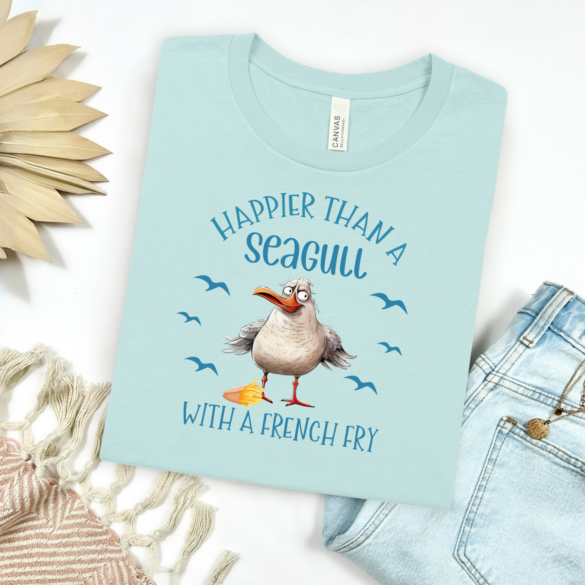 Bella and Canvas 3001 "Happier Than A Seagull With A French Fry" t shirt with an illustration of a seagull with a box of French fries and seagulls flying in the air. Picture on a flay lay mockup with pair of blue jeans and blanket and a wicker sun shaped accessory.