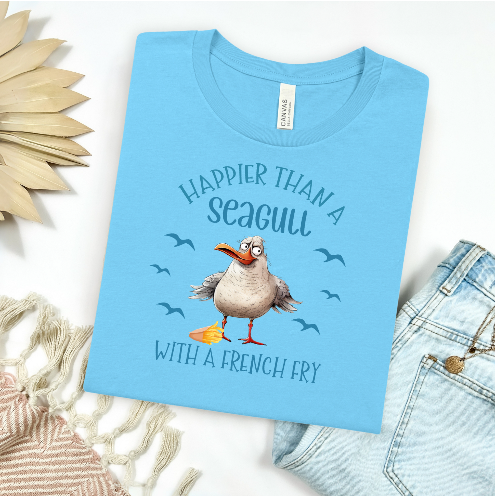 Bella and Canvas 3001 "Happier Than A Seagull With A French Fry" t shirt with an illustration of a seagull with a box of French fries and seagulls flying in the air. Picture on a flay lay mockup with pair of blue jeans and blanket and a wicker sun shaped accessory.