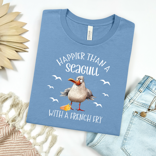Bella and Canvas 3001 "Happier Than A Seagull With A French Fry" t shirt with an illustration of a seagull with a box of French fries and seagulls flying in the air. Picture on a flay lay mockup with pair of blue jeans and blanket and a wicker sun shaped accessory.