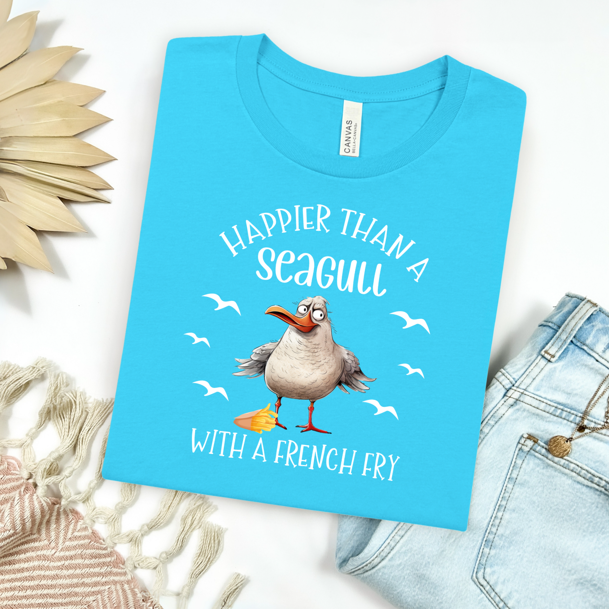 Bella and Canvas 3001 "Happier Than A Seagull With A French Fry" t shirt with an illustration of a seagull with a box of French fries and seagulls flying in the air. Picture on a flay lay mockup with pair of blue jeans and blanket and a wicker sun shaped accessory.