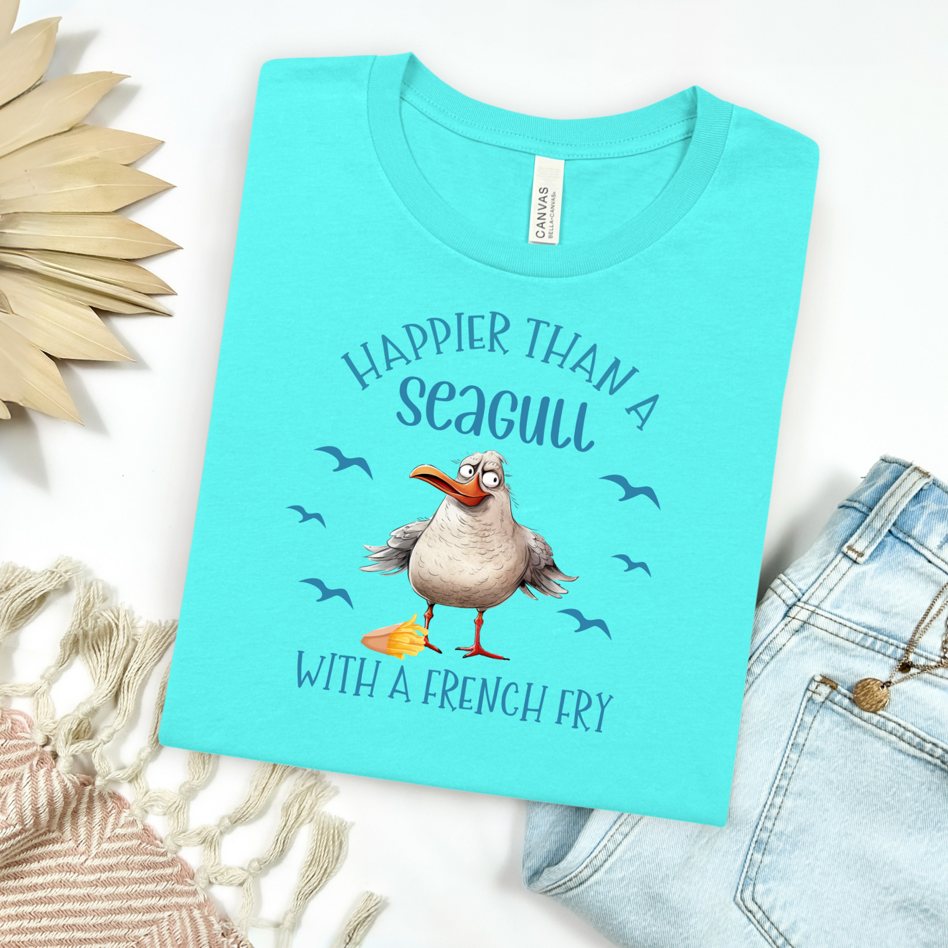Bella and Canvas 3001 "Happier Than A Seagull With A French Fry" t shirt with an illustration of a seagull with a box of French fries and seagulls flying in the air. Picture on a flay lay mockup with pair of blue jeans and blanket and a wicker sun shaped accessory.