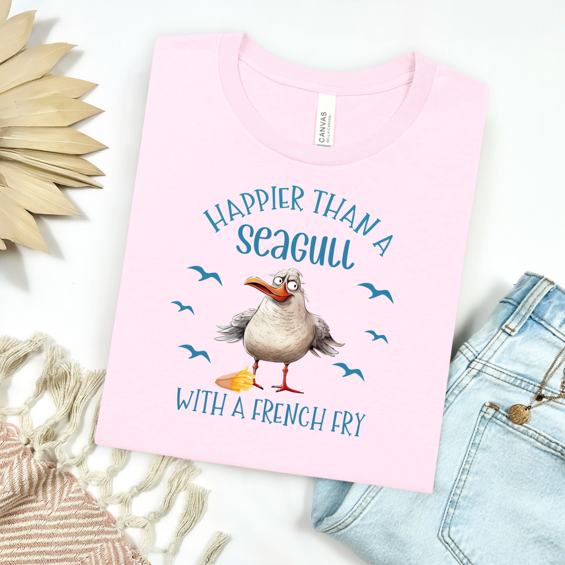 Bella and Canvas 3001 "Happier Than A Seagull With A French Fry" t shirt with an illustration of a seagull with a box of French fries and seagulls flying in the air. Picture on a flay lay mockup with pair of blue jeans and blanket and a wicker sun shaped accessory.