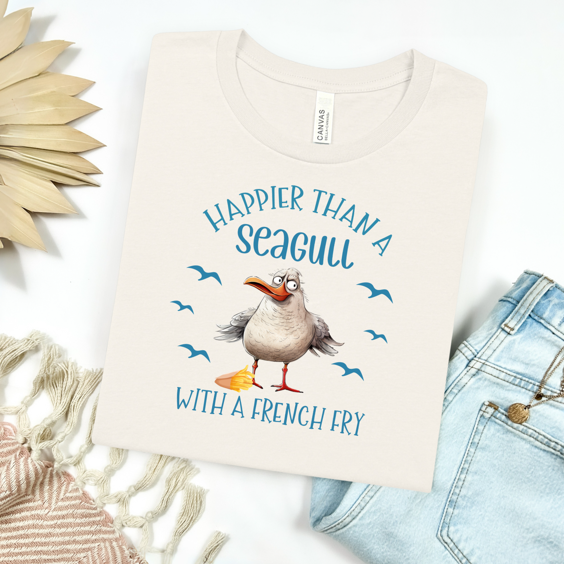 Bella and Canvas 3001 "Happier Than A Seagull With A French Fry" t shirt with an illustration of a seagull with a box of French fries and seagulls flying in the air. Picture on a flay lay mockup with pair of blue jeans and blanket and a wicker sun shaped accessory.