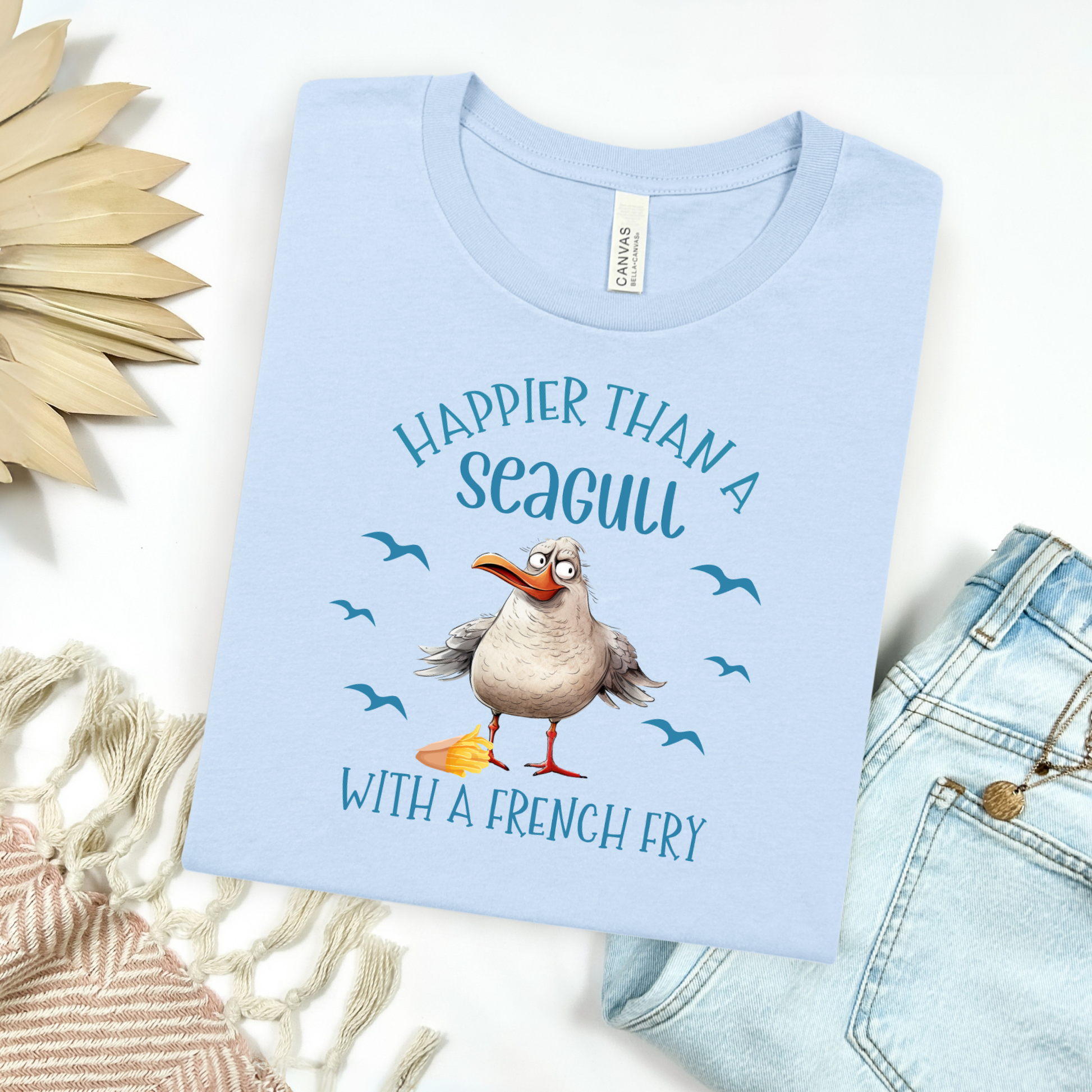 Bella and Canvas 3001 "Happier Than A Seagull With A French Fry" t shirt with an illustration of a seagull with a box of French fries and seagulls flying in the air. Picture on a flay lay mockup with pair of blue jeans and blanket and a wicker sun shaped accessory.