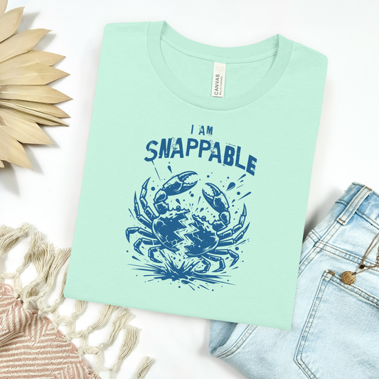 Bella Canvas 3001 "I Am Snappable" t shirt with a crab cracked in half in one color graphic. Picture on a flay lay mockup with pair of blue jeans and blanket and a wicker sun shaped accessory.