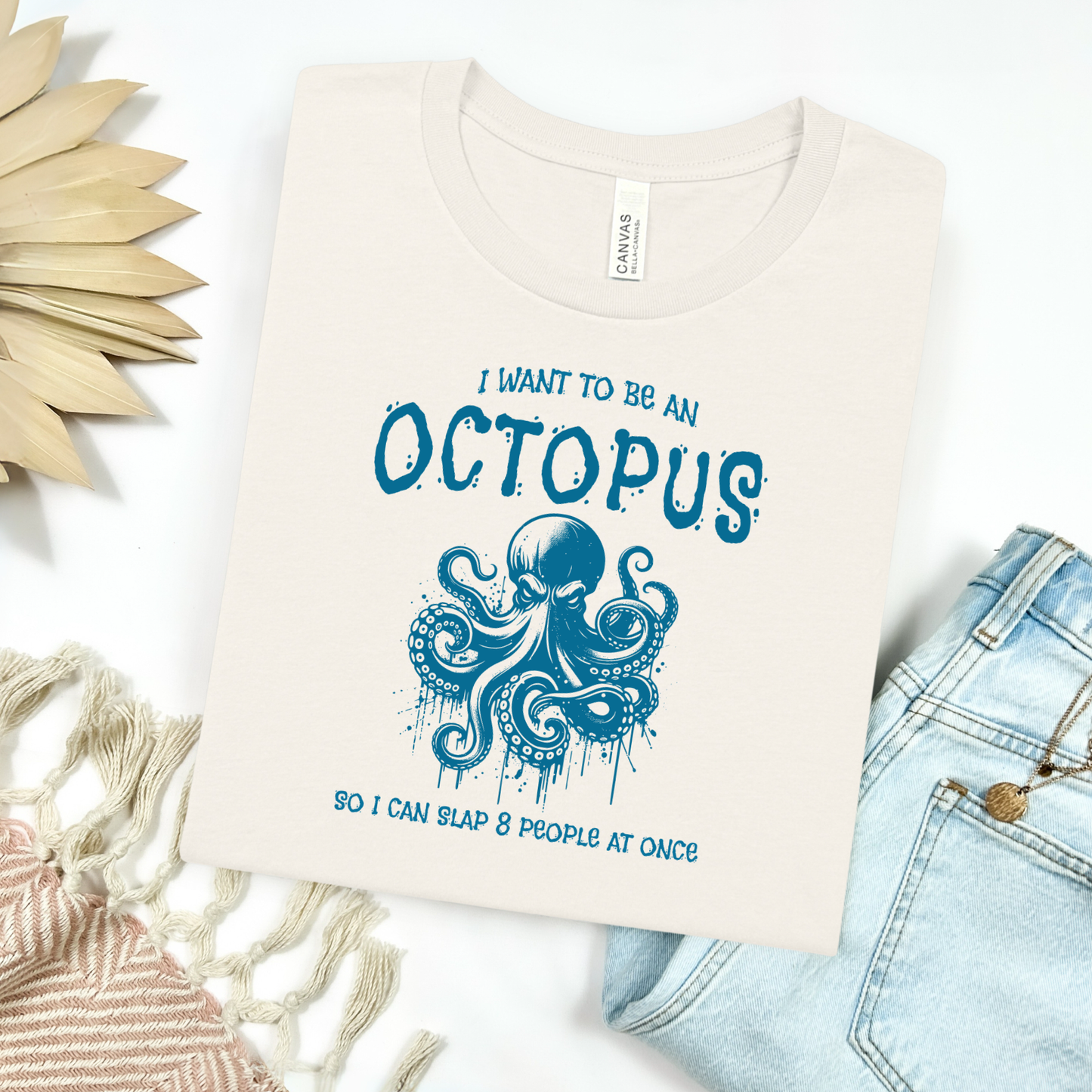 Bella Canvas 3001 "I Want To Be An Octopus So I Can Slap 8 People At Once" t shirt with an angry octopus one color graphic. Picture on a flay lay mockup with pair of blue jeans and blanket and a wicker sun shaped accessory.