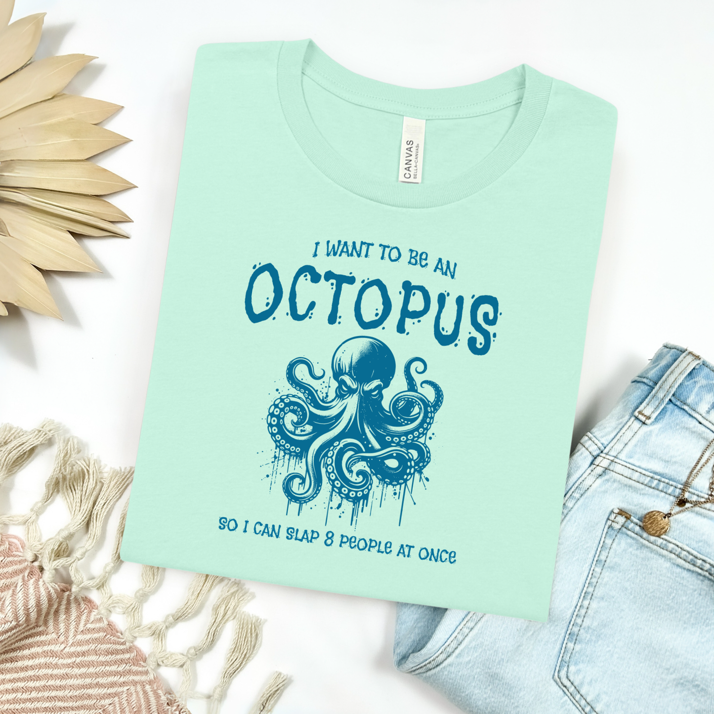 Bella Canvas 3001 "I Want To Be An Octopus So I Can Slap 8 People At Once" t shirt with an angry octopus one color graphic. Picture on a flay lay mockup with pair of blue jeans and blanket and a wicker sun shaped accessory.