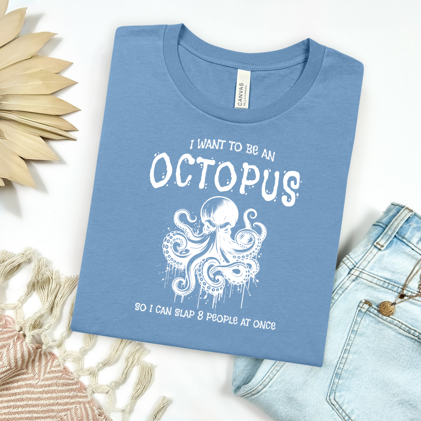 Bella Canvas 3001 "I Want To Be An Octopus So I Can Slap 8 People At Once" t shirt with an angry octopus one color graphic. Picture on a flay lay mockup with pair of blue jeans and blanket and a wicker sun shaped accessory.