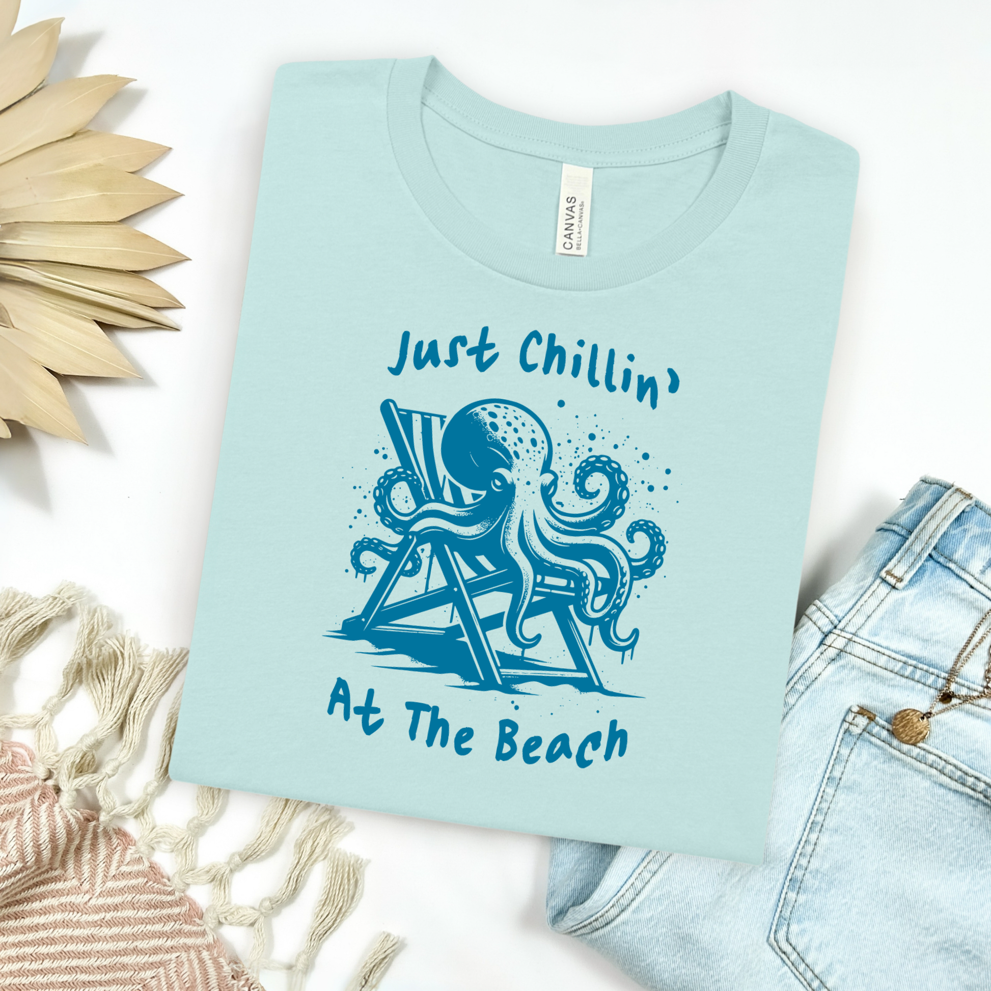 Bella Canvas 3001 "Just Chillin' At The Beach" t shirt with an octopus lounging in a beach chair in a one color graphic. Picture on a flay lay mockup with pair of blue jeans and blanket and a wicker sun shaped accessory.