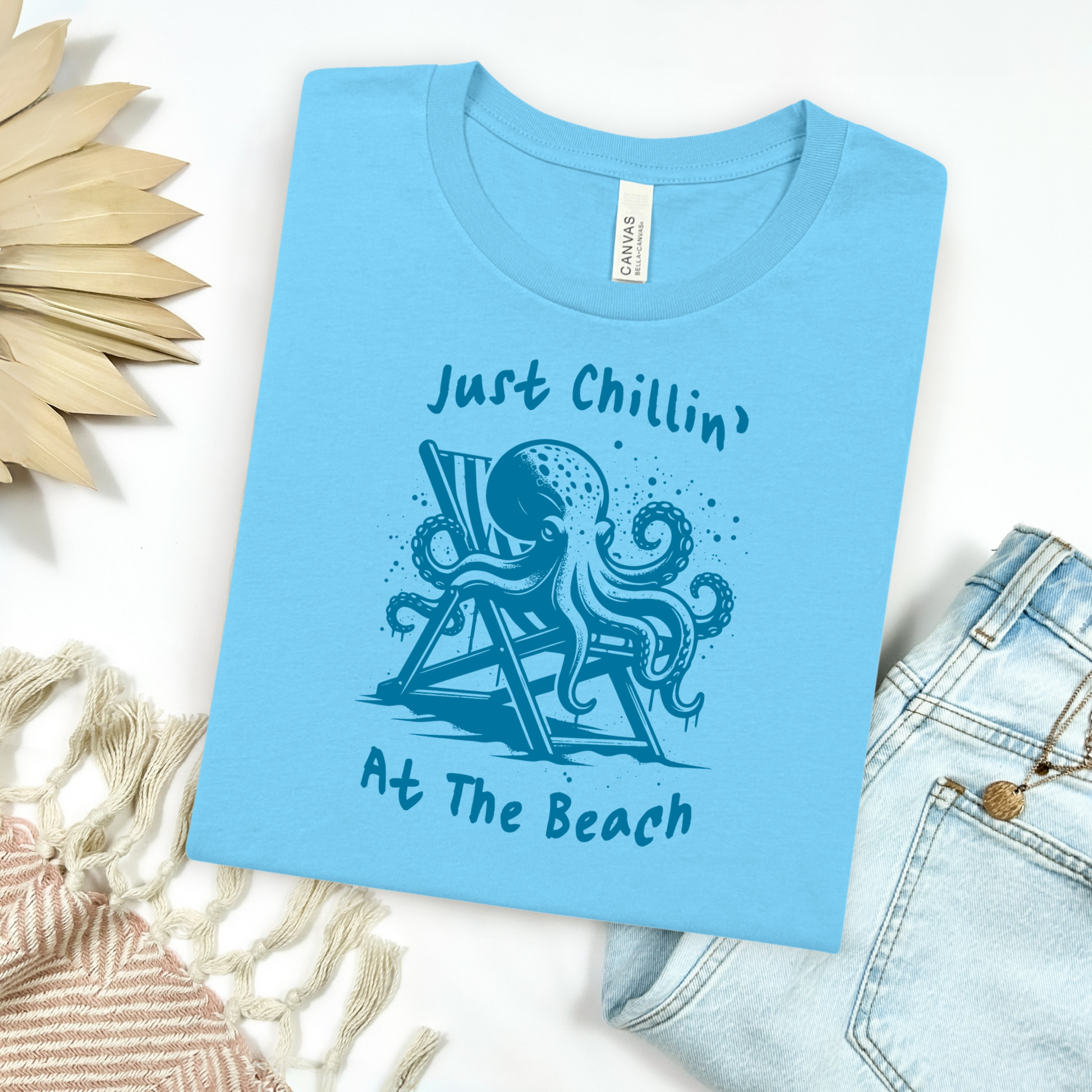 Bella Canvas 3001 "Just Chillin' At The Beach" t shirt with an octopus lounging in a beach chair in a one color graphic. Picture on a flay lay mockup with pair of blue jeans and blanket and a wicker sun shaped accessory.