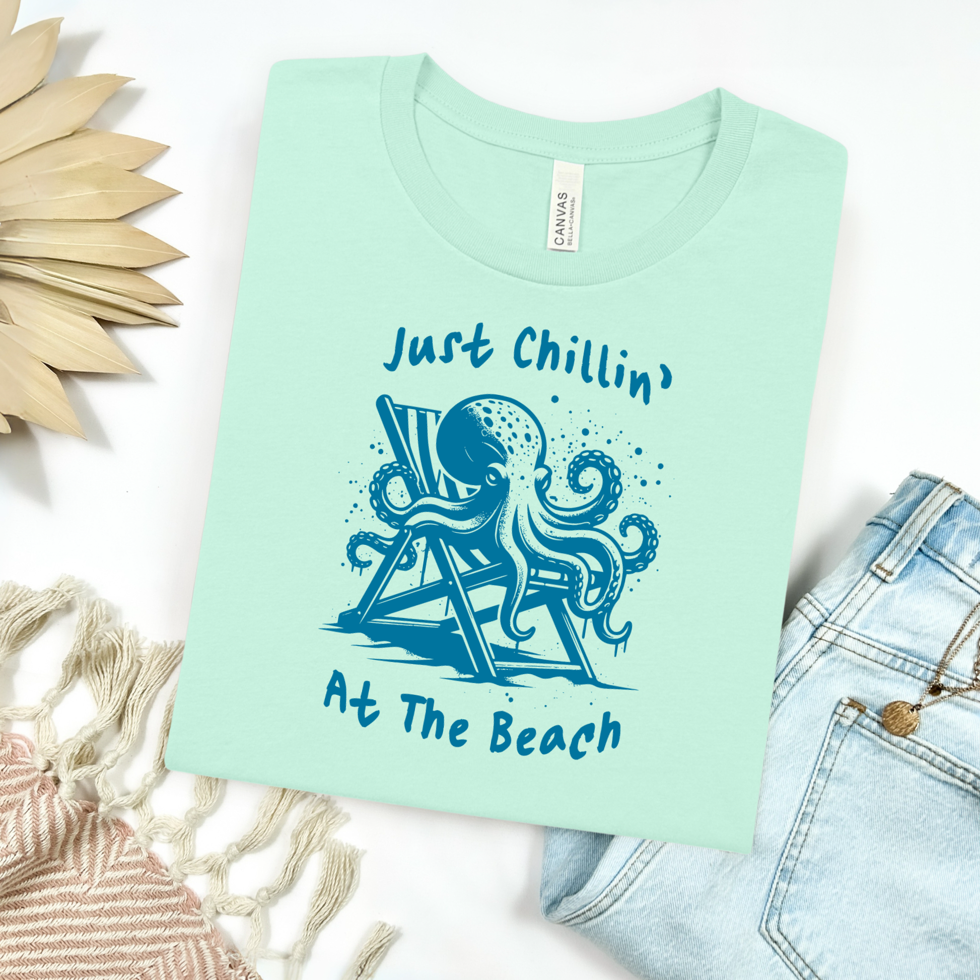 Bella Canvas 3001 "Just Chillin' At The Beach" t shirt with an octopus lounging in a beach chair in a one color graphic. Picture on a flay lay mockup with pair of blue jeans and blanket and a wicker sun shaped accessory.
