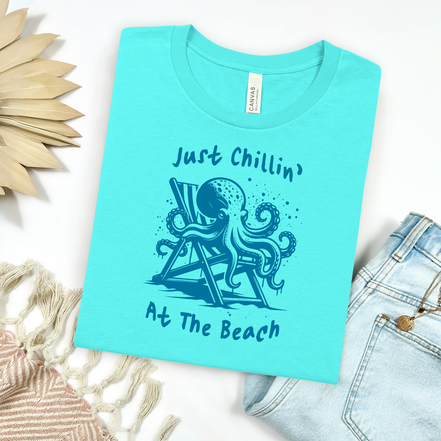 Bella Canvas 3001 "Just Chillin' At The Beach" t shirt with an octopus lounging in a beach chair in a one color graphic. Picture on a flay lay mockup with pair of blue jeans and blanket and a wicker sun shaped accessory.