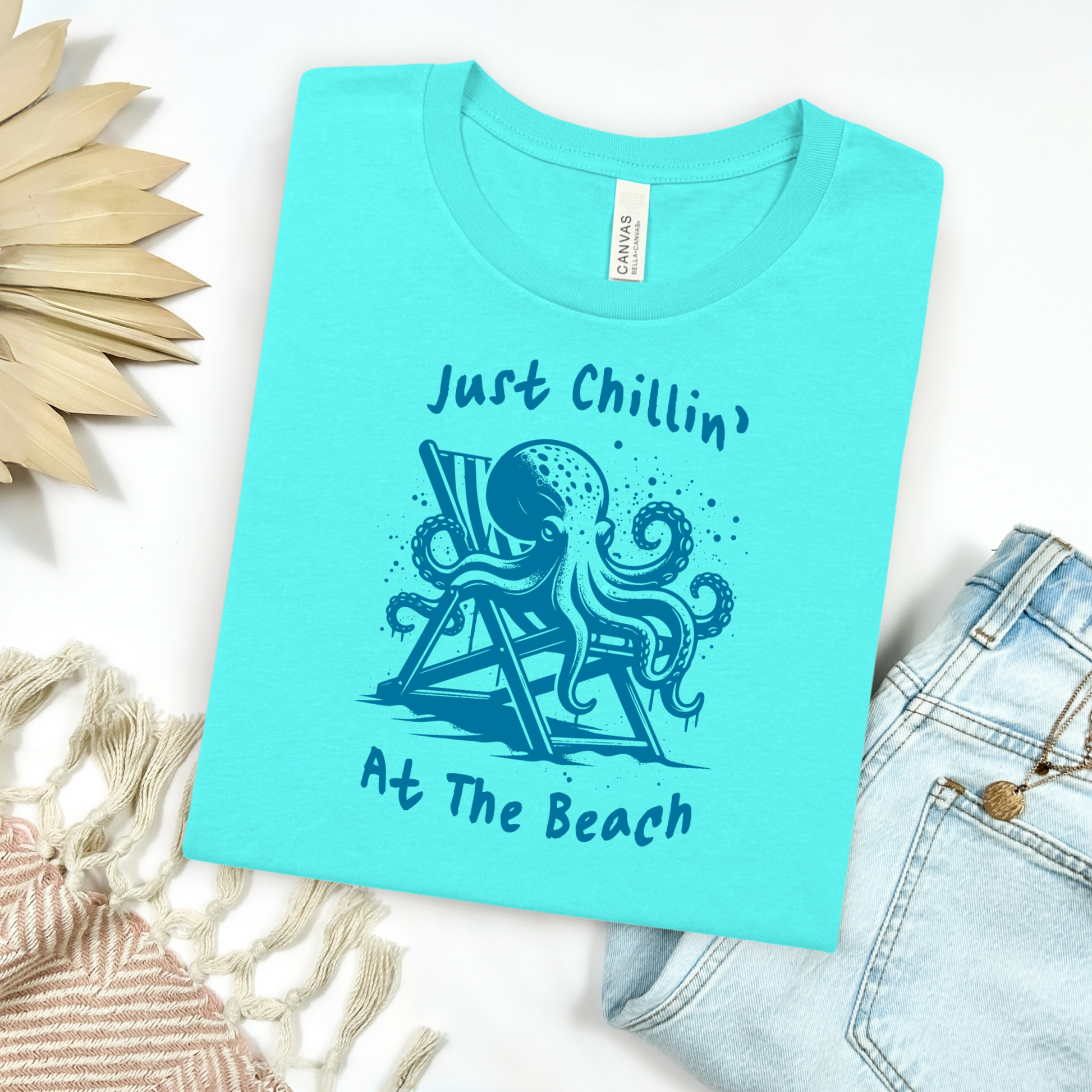 Bella Canvas 3001 "Just Chillin' At The Beach" t shirt with an octopus lounging in a beach chair in a one color graphic. Picture on a flay lay mockup with pair of blue jeans and blanket and a wicker sun shaped accessory.