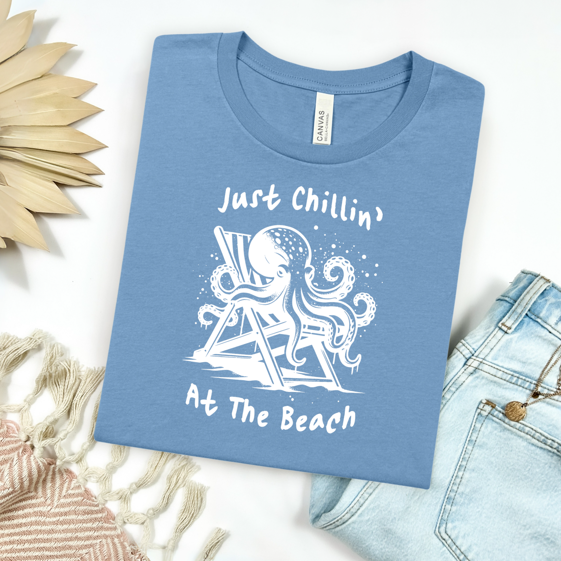 Bella Canvas 3001 "Just Chillin' At The Beach" t shirt with an octopus lounging in a beach chair in a one color graphic. Picture on a flay lay mockup with pair of blue jeans and blanket and a wicker sun shaped accessory.