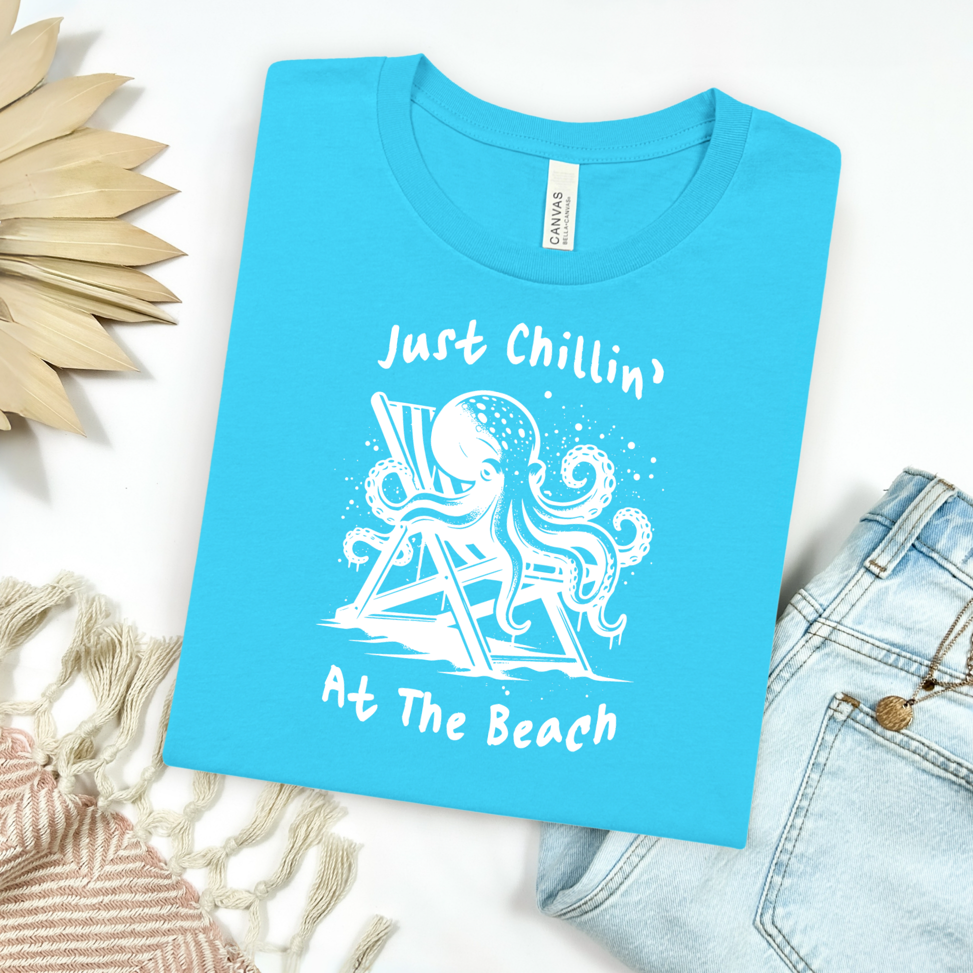 Bella Canvas 3001 "Just Chillin' At The Beach" t shirt with an octopus lounging in a beach chair in a one color graphic. Picture on a flay lay mockup with pair of blue jeans and blanket and a wicker sun shaped accessory.