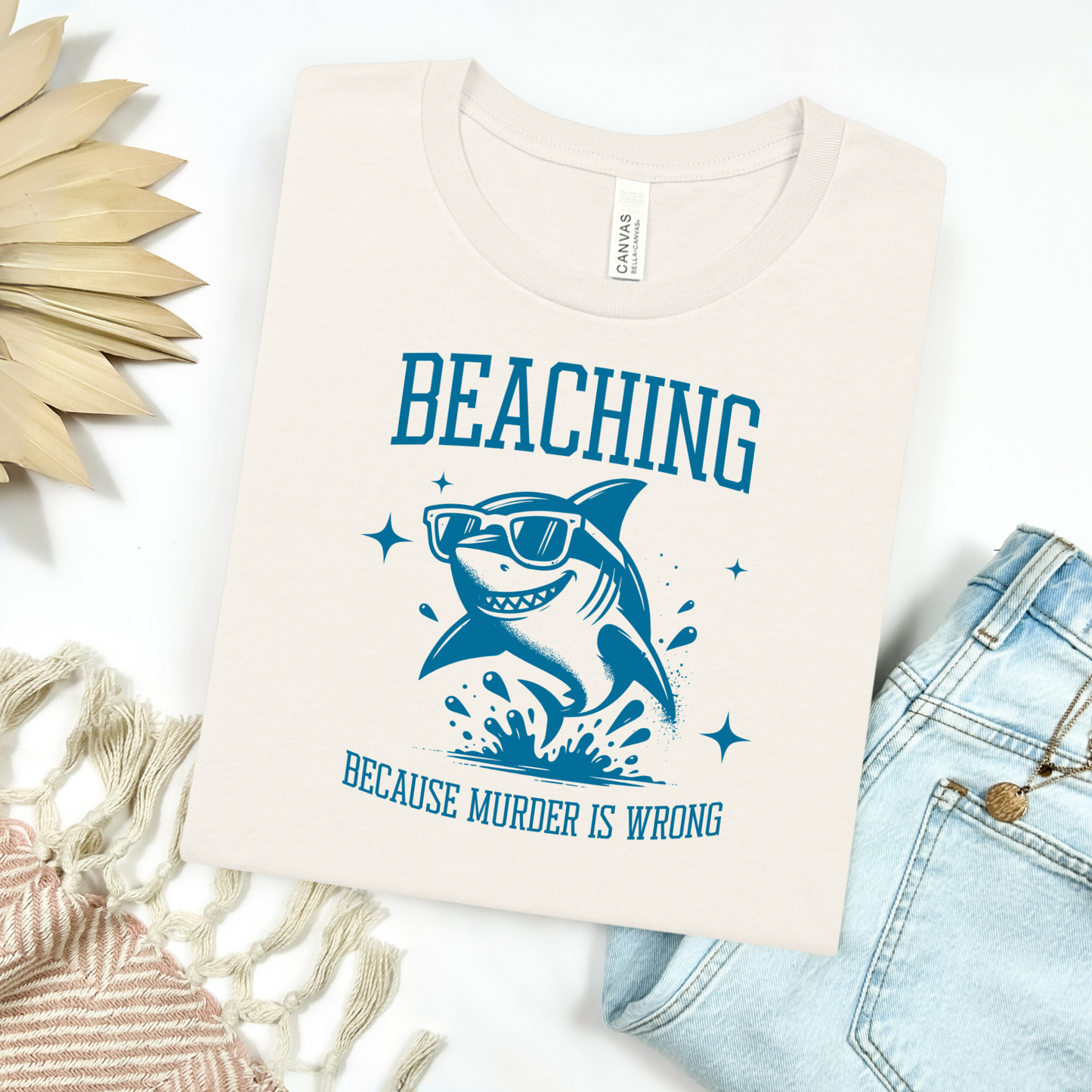 Bella Canvas 3001 Beaching Because Murder Is Wrong  T Shirt with Funny Shark with Sunglasses splashing in water in one color graphic. Picture on a flay lay mockup with pair of blue jeans and blanket and a wicker sun shaped accessory.