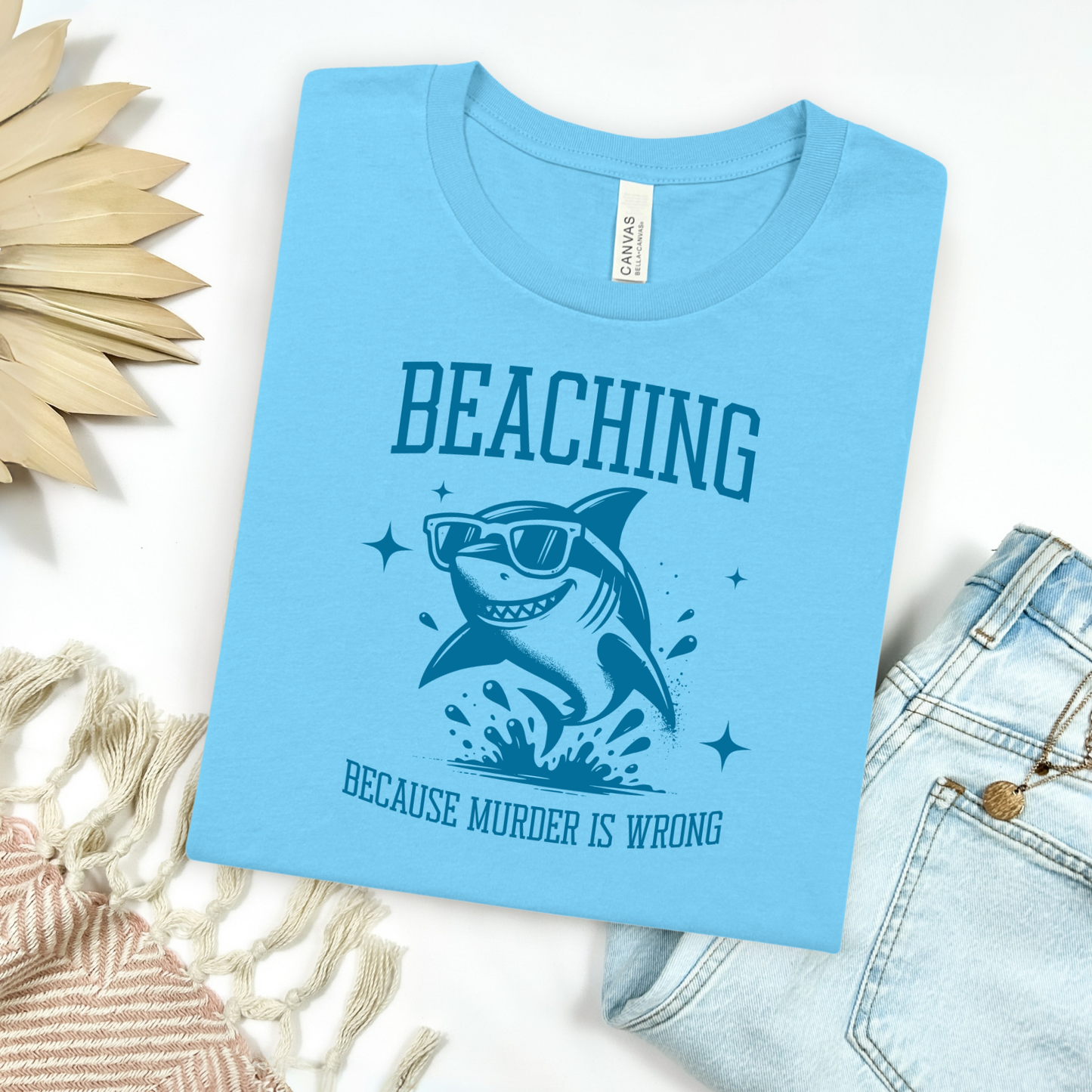 Bella Canvas 3001 Beaching Because Murder Is Wrong  T Shirt with Funny Shark with Sunglasses splashing in water in one color graphic. Picture on a flay lay mockup with pair of blue jeans and blanket and a wicker sun shaped accessory.