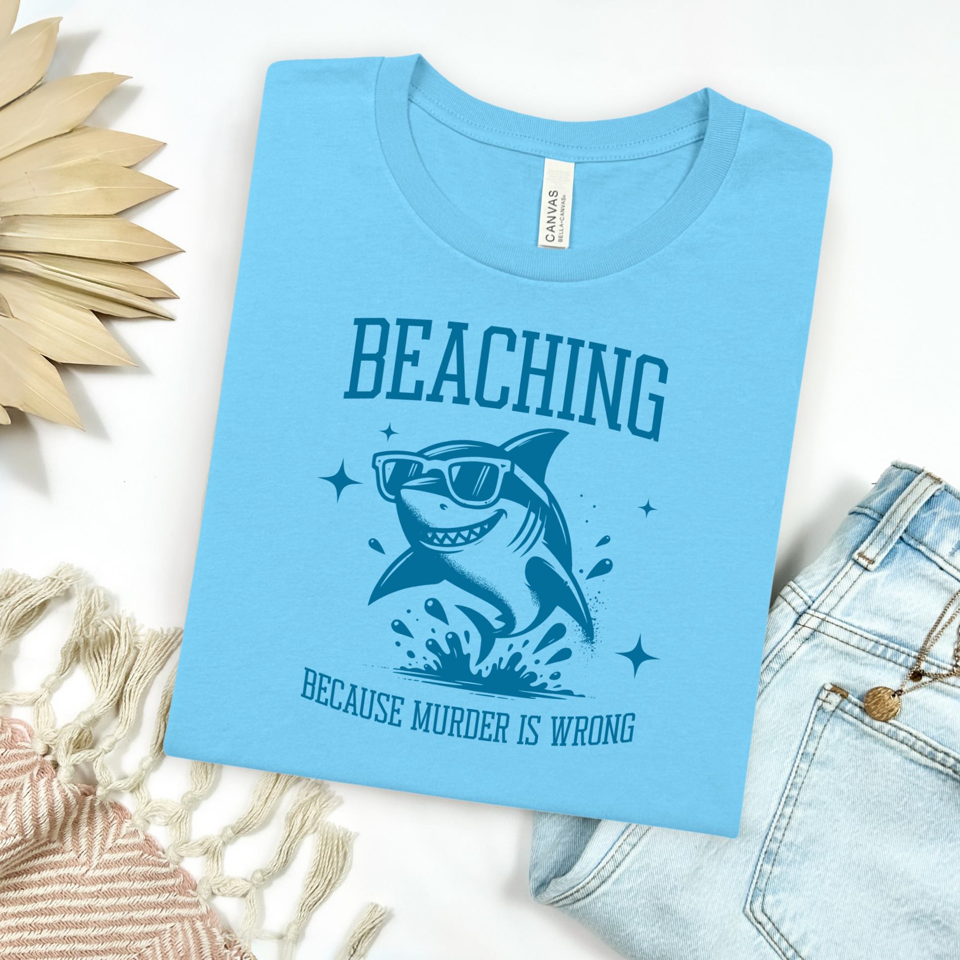 Bella Canvas 3001 Beaching Because Murder Is Wrong  T Shirt with Funny Shark with Sunglasses splashing in water in one color graphic. Picture on a flay lay mockup with pair of blue jeans and blanket and a wicker sun shaped accessory.
