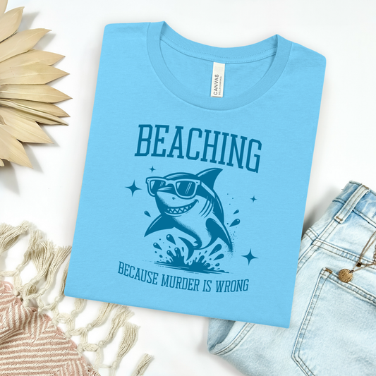 Bella Canvas 3001 Beaching Because Murder Is Wrong  T Shirt with Funny Shark with Sunglasses splashing in water in one color graphic. Picture on a flay lay mockup with pair of blue jeans and blanket and a wicker sun shaped accessory.