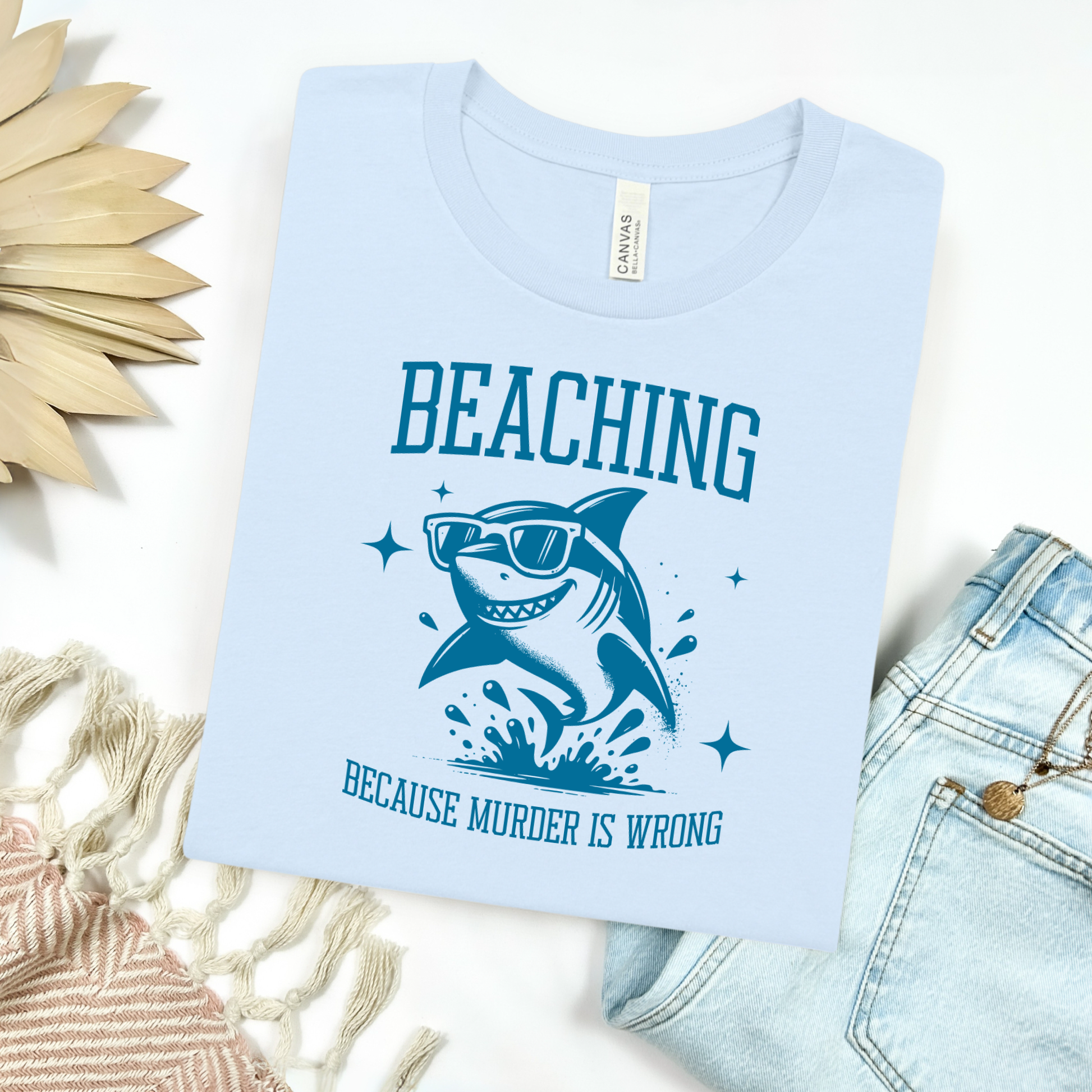Bella Canvas 3001 Beaching Because Murder Is Wrong  T Shirt with Funny Shark with Sunglasses splashing in water in one color graphic. Picture on a flay lay mockup with pair of blue jeans and blanket and a wicker sun shaped accessory.