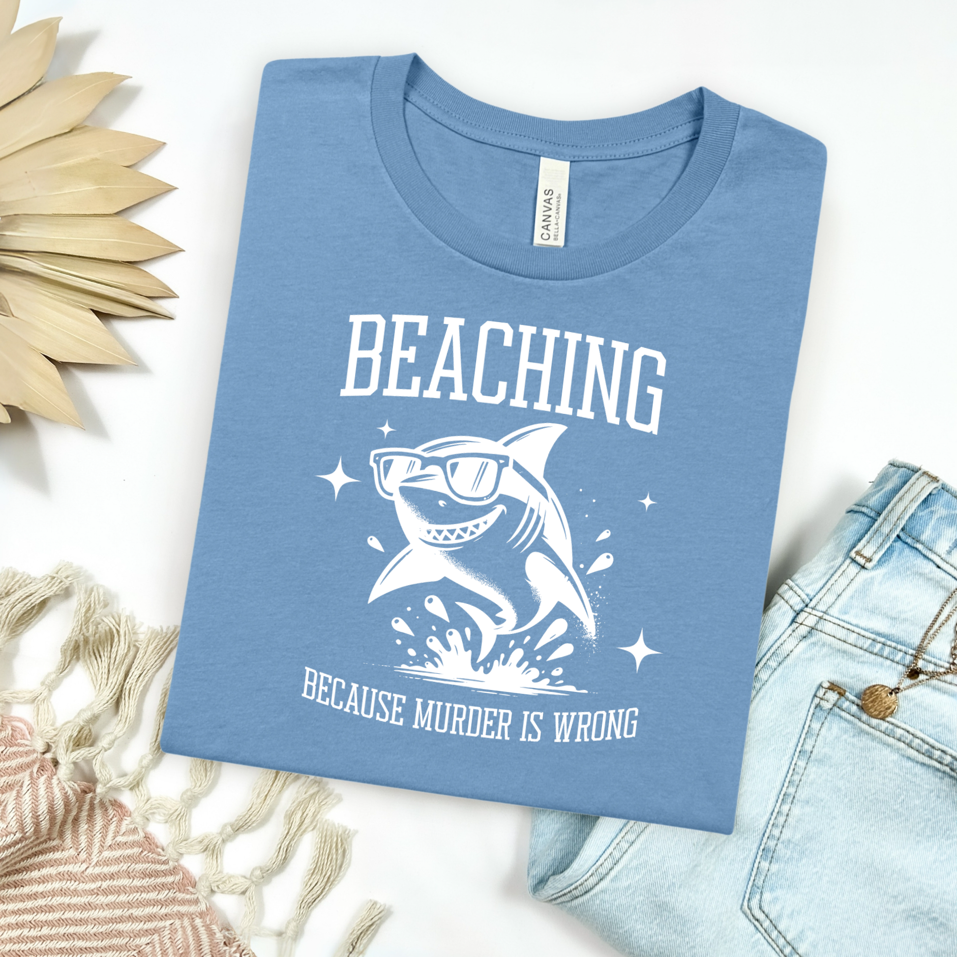 Bella Canvas 3001 Beaching Because Murder Is Wrong  T Shirt with Funny Shark with Sunglasses splashing in water in one color graphic. Picture on a flay lay mockup with pair of blue jeans and blanket and a wicker sun shaped accessory.
