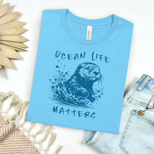 Bella Canvas 3001 " Save The Ocean And The Sea Life In It" t shirt of a sea otter splashing in water in one color graphic. Picture on a flay lay mockup with pair of blue jeans and blanket and a wicker sun shaped accessory.