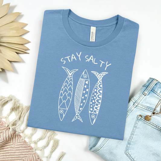 Bella Canvas 3001 "Stay Salty" t shirt with three artsy style sardines one color graphic. Picture on a flay lay mockup with pair of blue jeans and blanket and a wicker sun shaped accessory.