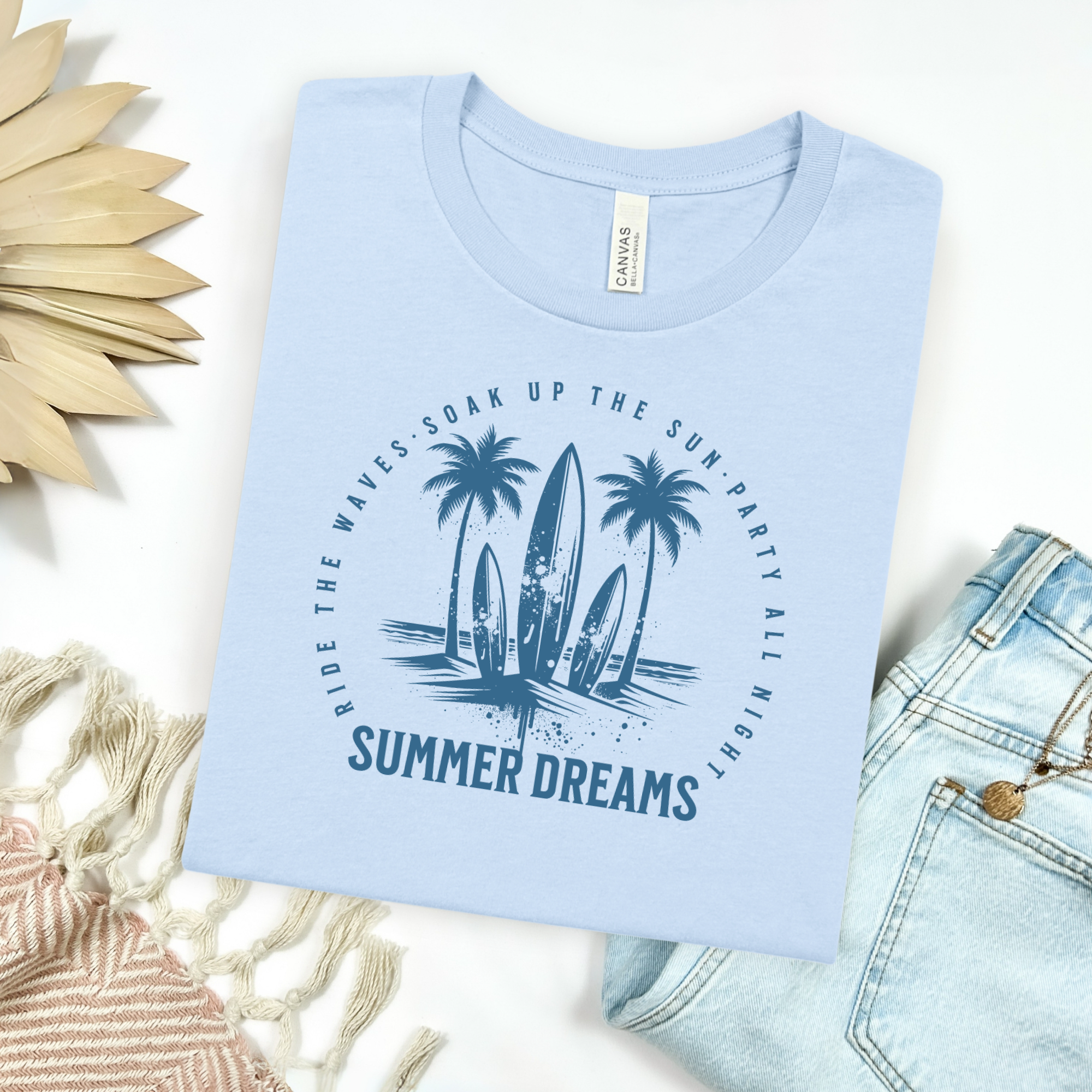 Bella canvas 3001 "Summer Dreams" t shirt with surfboards in center with inspiring text around the graphic in one color graphic. Picture on a flay lay mockup with pair of blue jeans and blanket and a wicker sun shaped accessory.