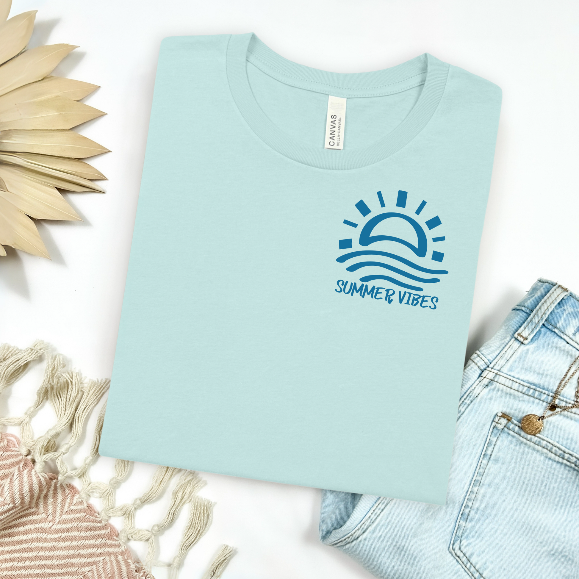 Bella and Canvas 3001 "Summer Vibes" t shirt with sun and waves one color graphic. Picture on a flay lay mockup with pair of blue jeans and blanket and a wicker sun shaped accessory.