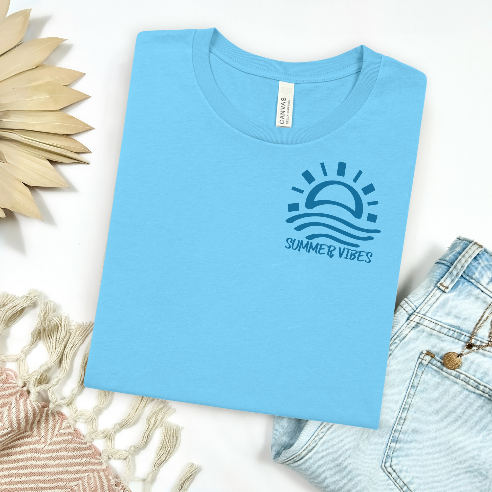 Bella and Canvas 3001 "Summer Vibes" t shirt with sun and waves one color graphic. Picture on a flay lay mockup with pair of blue jeans and blanket and a wicker sun shaped accessory.