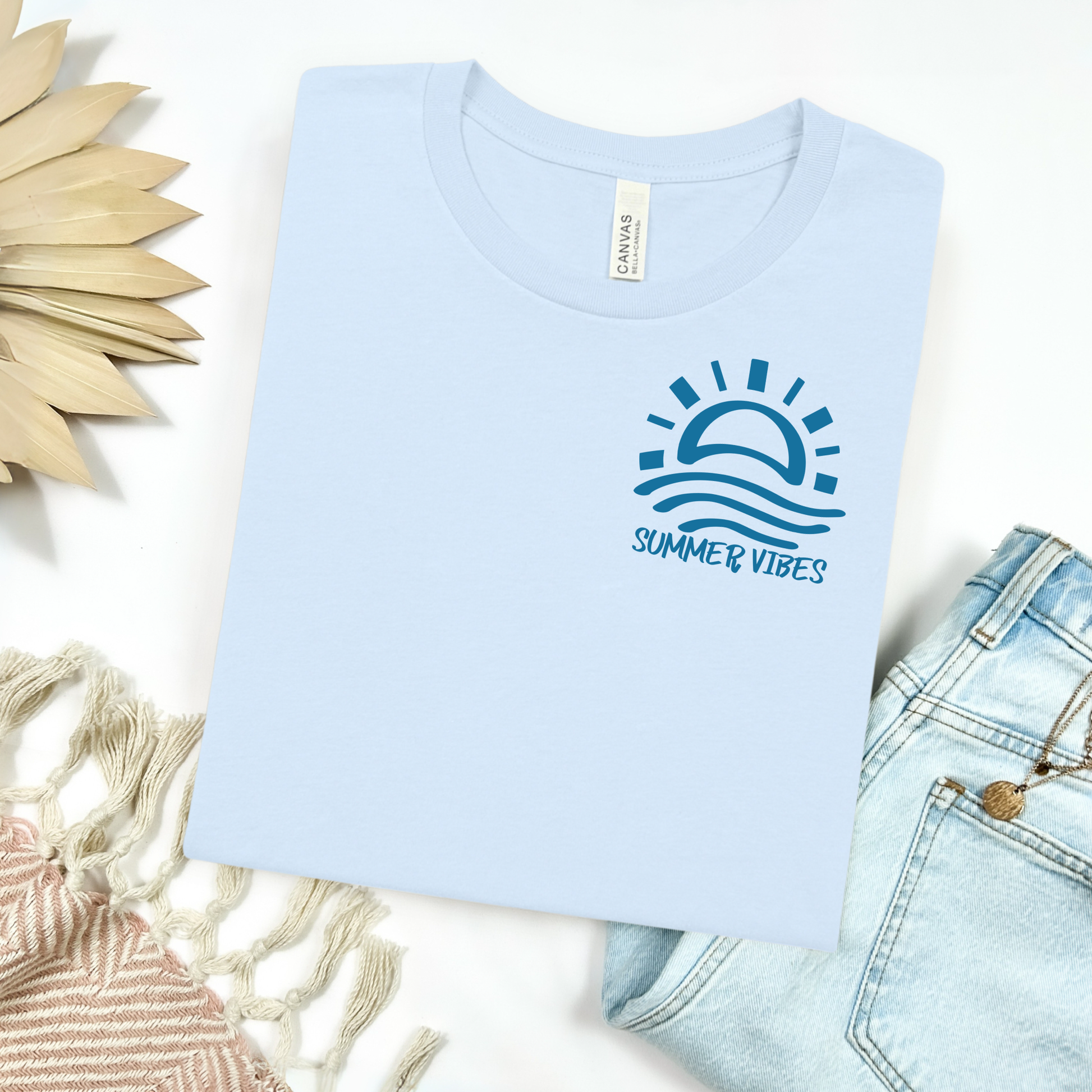 Bella and Canvas 3001 "Summer Vibes" t shirt with sun and waves one color graphic. Picture on a flay lay mockup with pair of blue jeans and blanket and a wicker sun shaped accessory.