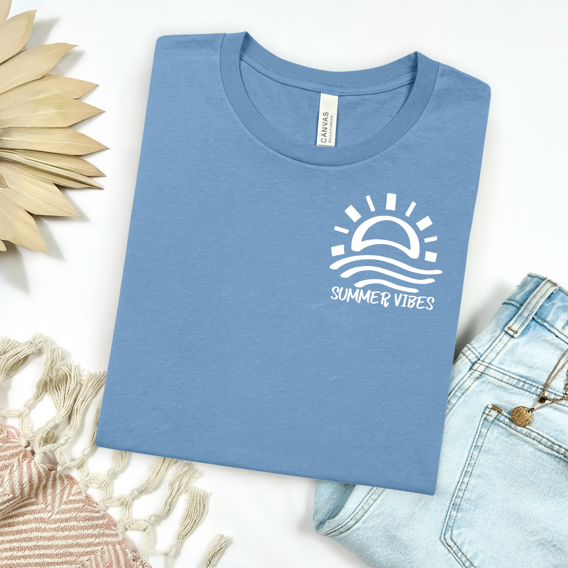 Bella and Canvas 3001 "Summer Vibes" t shirt with sun and waves one color graphic. Picture on a flay lay mockup with pair of blue jeans and blanket and a wicker sun shaped accessory.