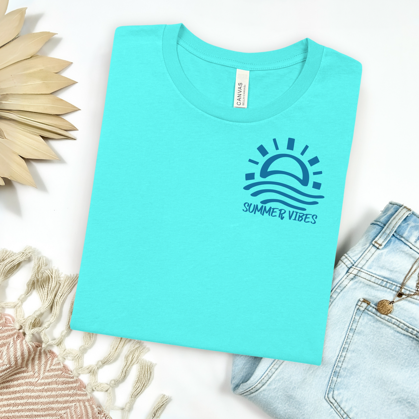 Bella and Canvas 3001 "Summer Vibes" t shirt with sun and waves one color graphic. Picture on a flay lay mockup with pair of blue jeans and blanket and a wicker sun shaped accessory.