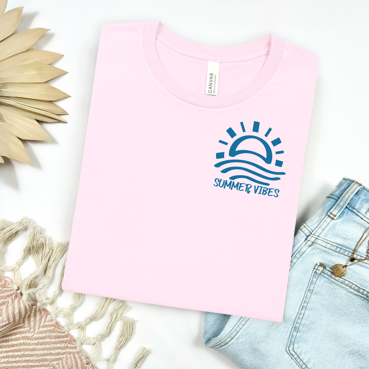 Bella and Canvas 3001 "Summer Vibes" t shirt with sun and waves one color graphic. Picture on a flay lay mockup with pair of blue jeans and blanket and a wicker sun shaped accessory.