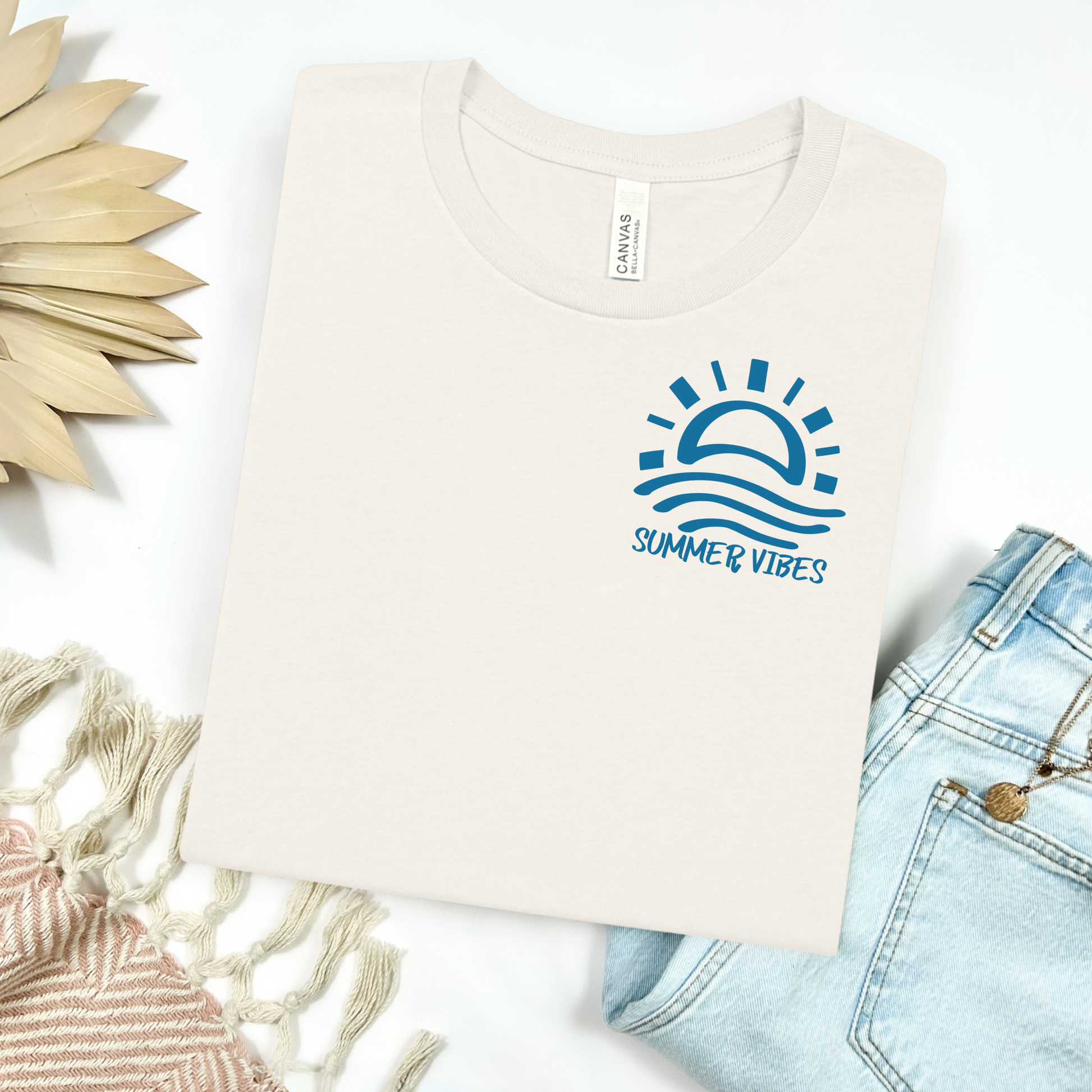 Bella and Canvas 3001 "Summer Vibes" t shirt with sun and waves one color graphic. Picture on a flay lay mockup with pair of blue jeans and blanket and a wicker sun shaped accessory.